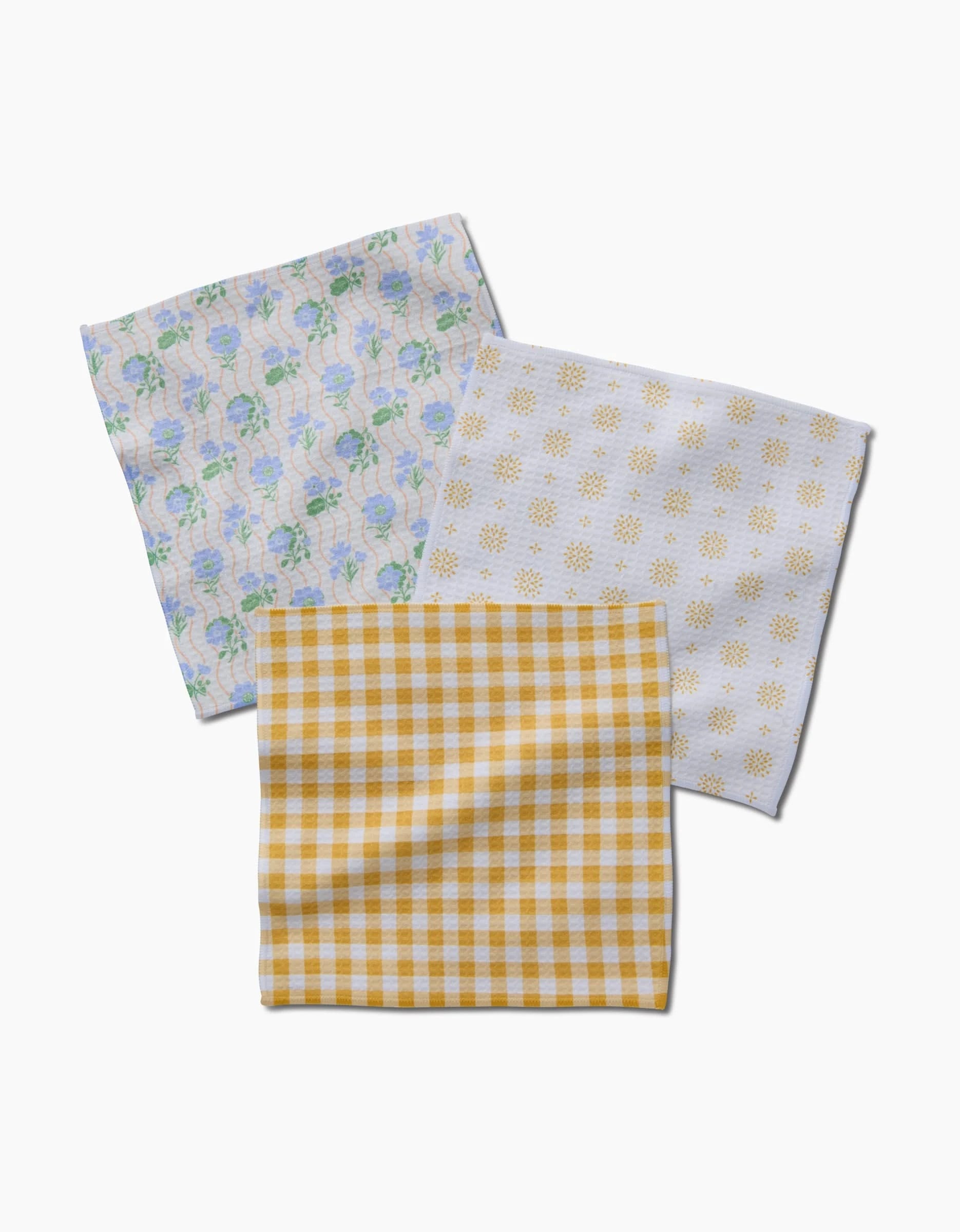 Three folded Danelys Sidron Spring Morning cloth napkins—blue and green floral, yellow sunburst, and yellow-white checkered—rest on a white surface, each crafted with ultra-absorbent Geoweave technology for a luxe feel.