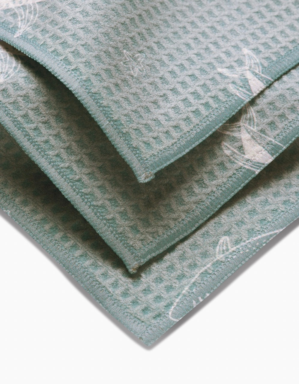 A close-up of three folded, teal-colored Toothy Tales washcloths by Janna Sue Design, featuring a waffle texture, subtle white sailboat designs, and crafted from recycled materials on a plain white background.