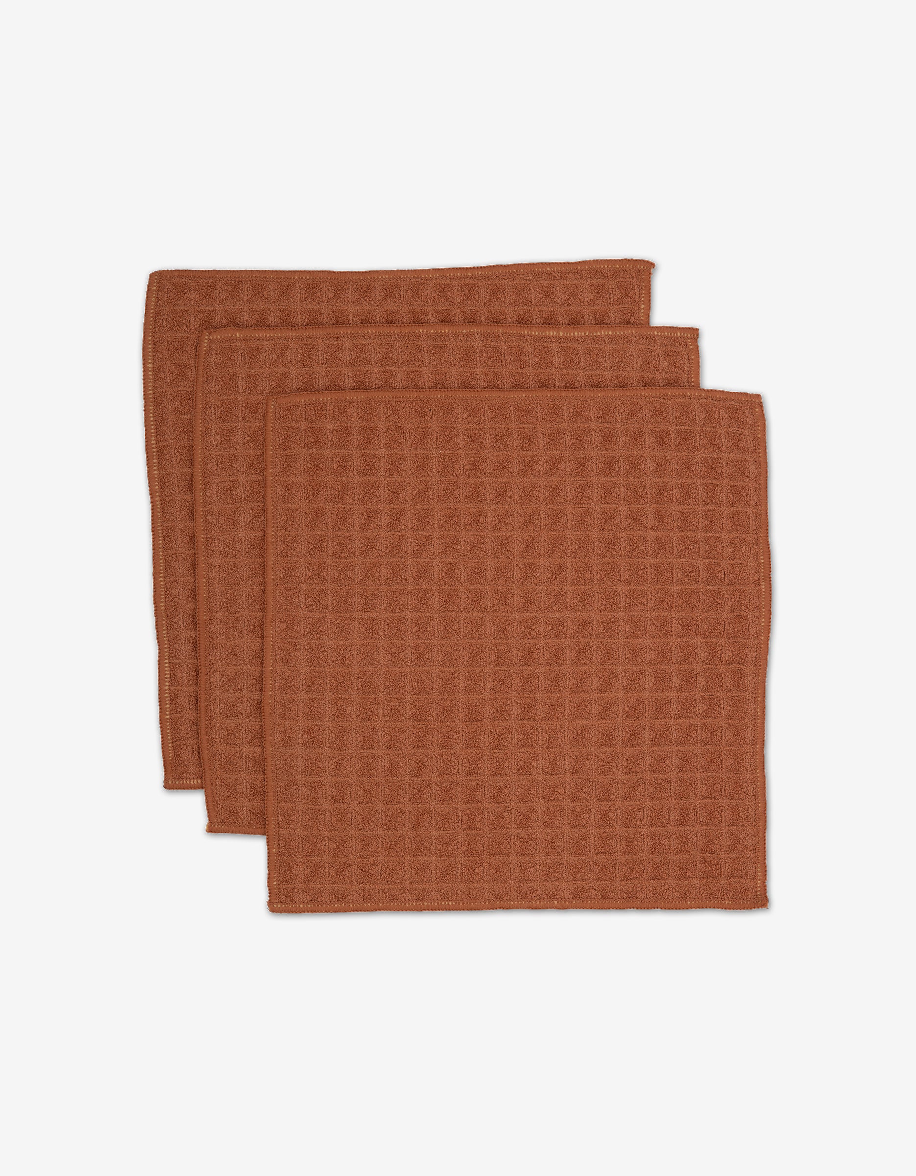 Three folded brown waffle-textured absorbent washcloths, stacked and slightly overlapping on a white background. GEOMETRY’s Clay washcloths from the Waffle Bath Collection.