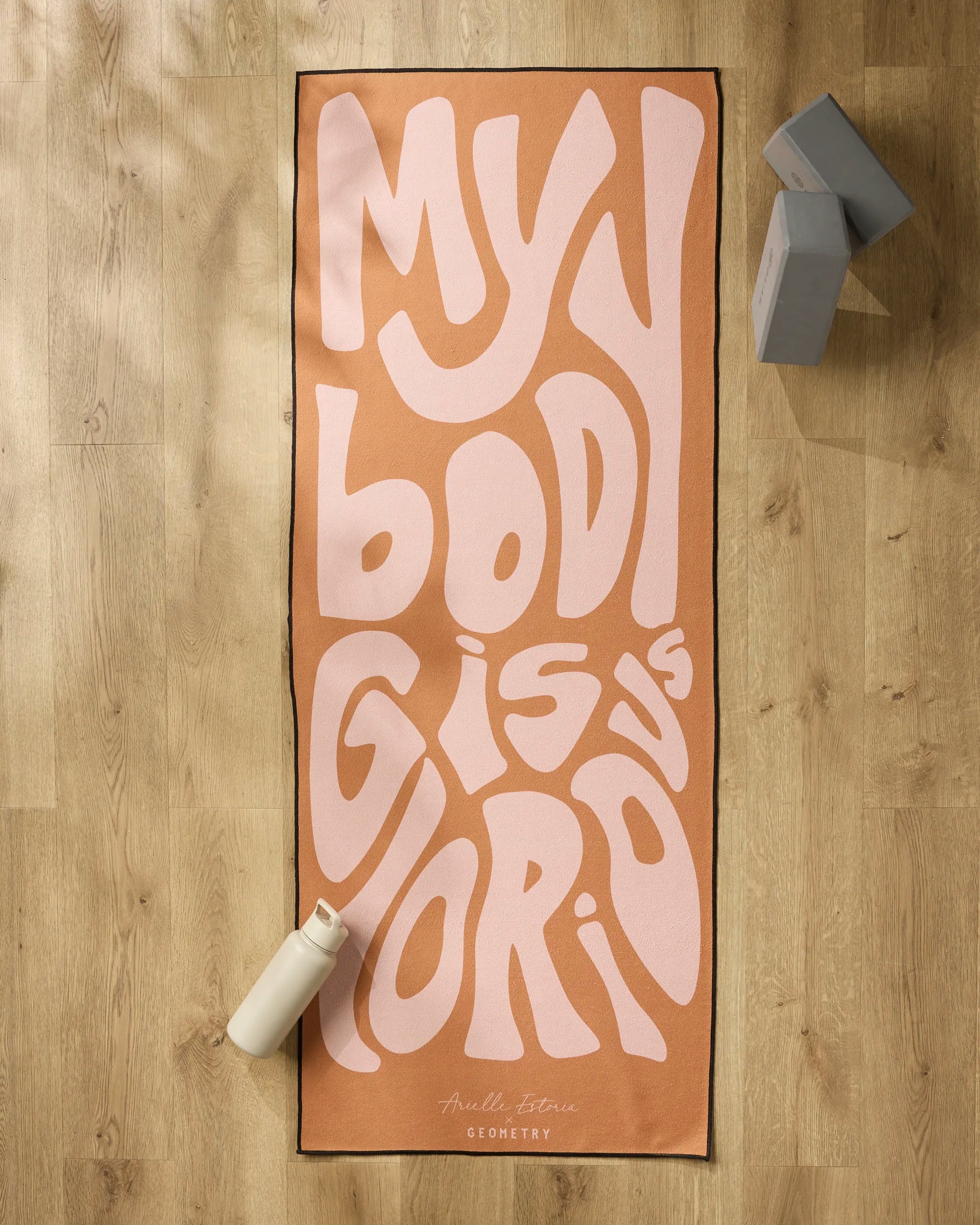 The peach-colored "Glorious" yoga mat by Arielle Estoria, featuring bold wavy pink MY BODY IS GLORIOUS text, is displayed on a light wood floor with a white water bottle, gray yoga blocks, and a non-slip towel for comfort and stability.