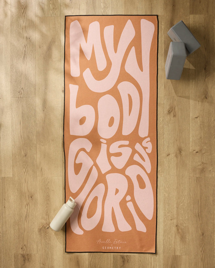 The peach-colored "Glorious" yoga mat by Arielle Estoria, featuring bold wavy pink MY BODY IS GLORIOUS text, is displayed on a light wood floor with a white water bottle, gray yoga blocks, and a non-slip towel for comfort and stability.