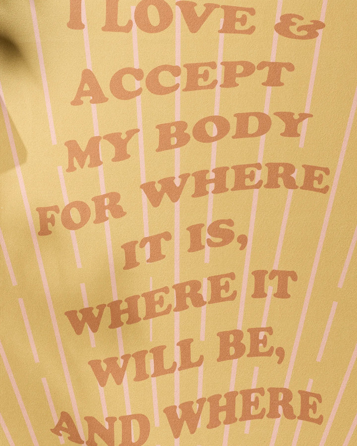 With a yellow background and pink lines, the "Love and Acceptance Affirmation" yoga towel by Arielle Estoria features bold text: I LOVE & ACCEPT MY BODY FOR WHERE IT IS, WHERE IT WILL BE, AND WHERE. Made from recycled materials to support self-love.