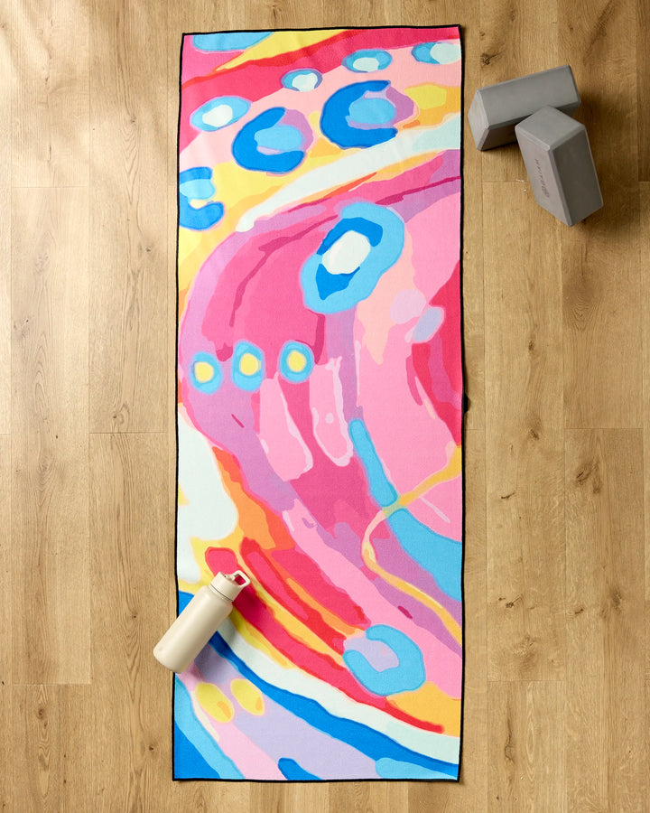 The "Abundance" recycled yoga mat towel by Sareka Unique, featuring vibrant pink, blue, yellow, and white abstract patterns, rests on a wooden floor beside two gray yoga blocks and a white water bottle with a pink lid.