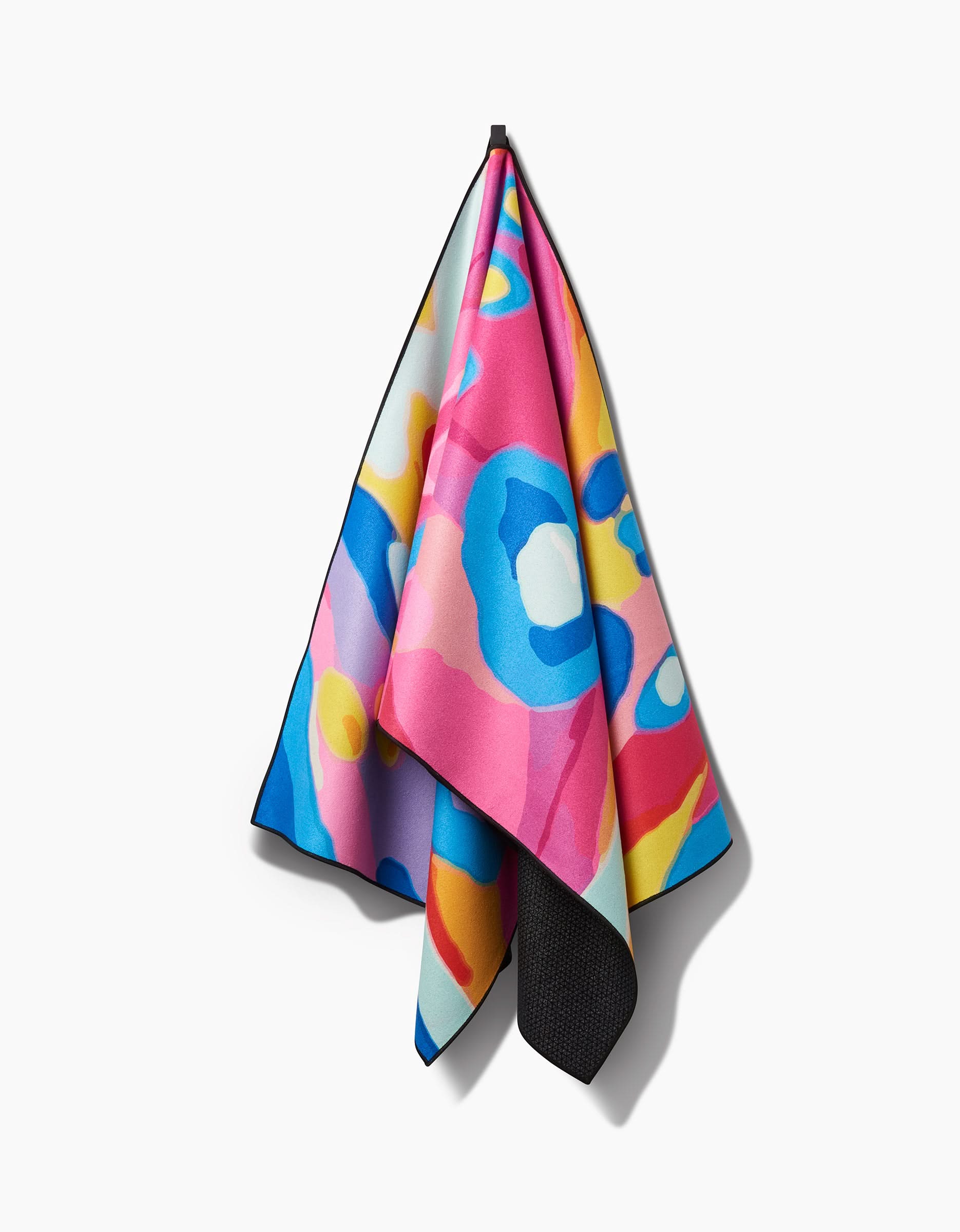 The Abundance yoga towel by Sareka Unique features a vibrant abstract pattern in pink, blue, yellow, and white. Made from recycled materials, it hangs from a single point against a plain white background.