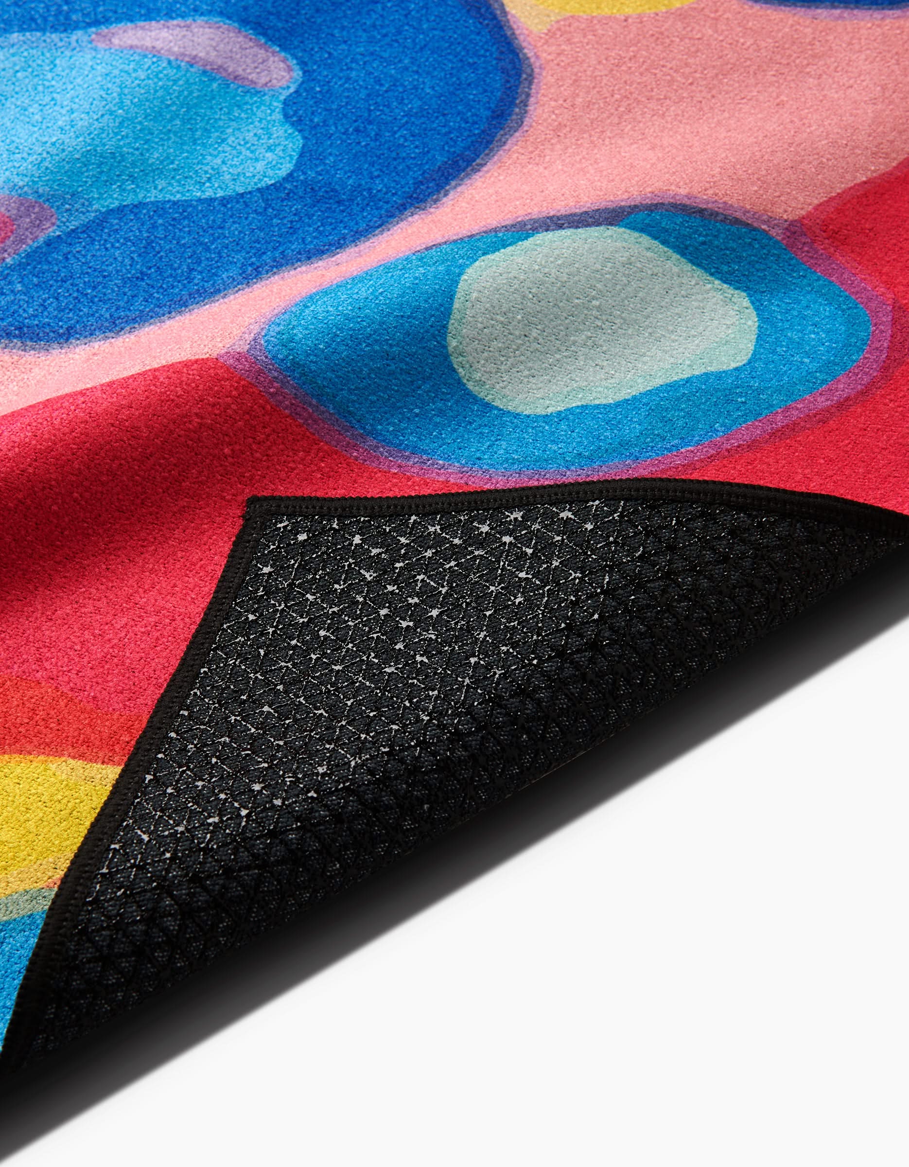 A close-up of the Sareka Unique Abundance rug, featuring abstract blue, pink, and red shapes and a black textured silicone anti-slip backing, slightly lifted at the corner.