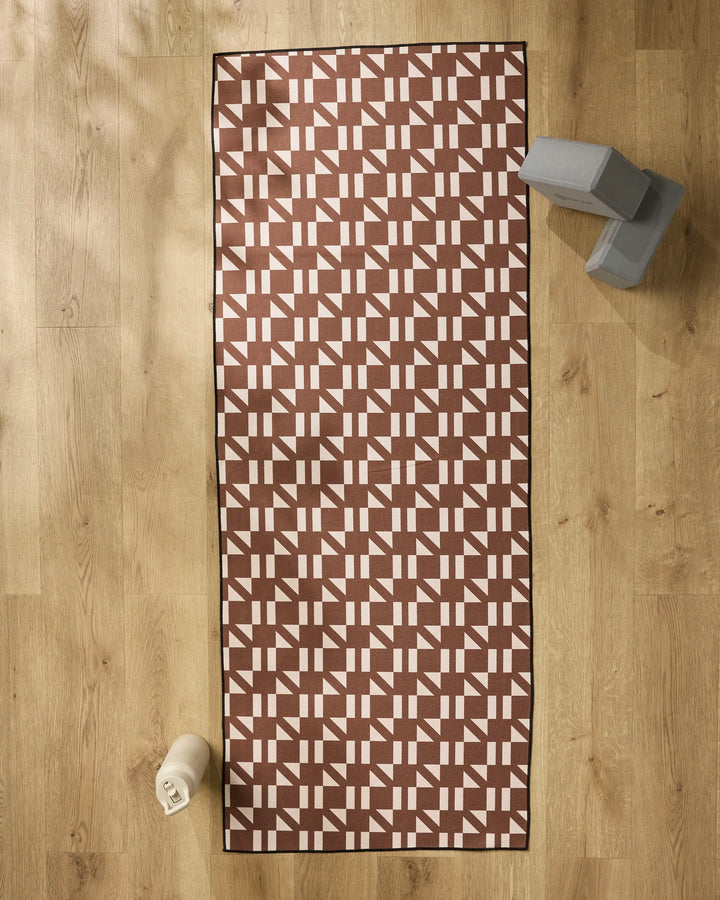 The Affogato yoga mat by Geometry, featuring a white geometric pattern on brown, is laid out on a light wooden floor. Nearby are gray yoga blocks, a water bottle, and a slip-resistant yoga towel for a calming workout setup.