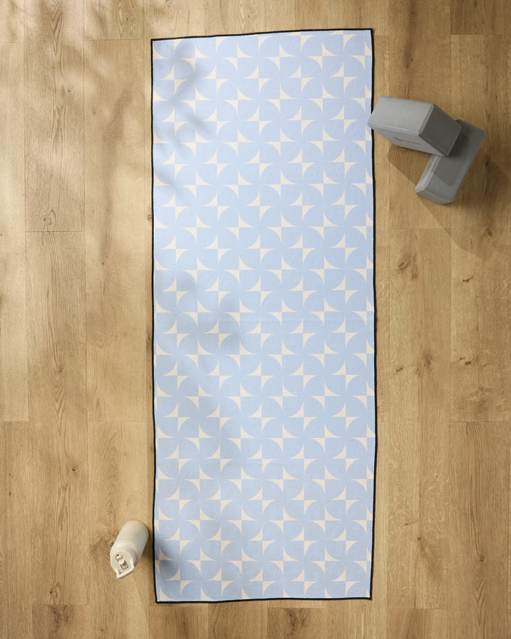 The Airy Flow yoga mat by Geometry, featuring a light blue color with a white geometric design, rests on a wooden floor with a silicone grip towel on top. A white water bottle sits bottom left; two gray yoga blocks are stacked top right.