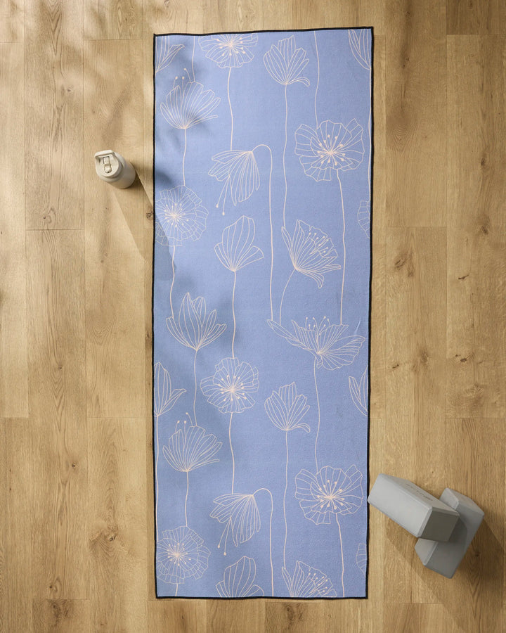 The Alexa by GEOMETRY, a blue yoga mat with white floral lines, is spread on a wooden floor and topped with a soft non-slip towel. A beige water bottle and two gray yoga blocks rest nearby.