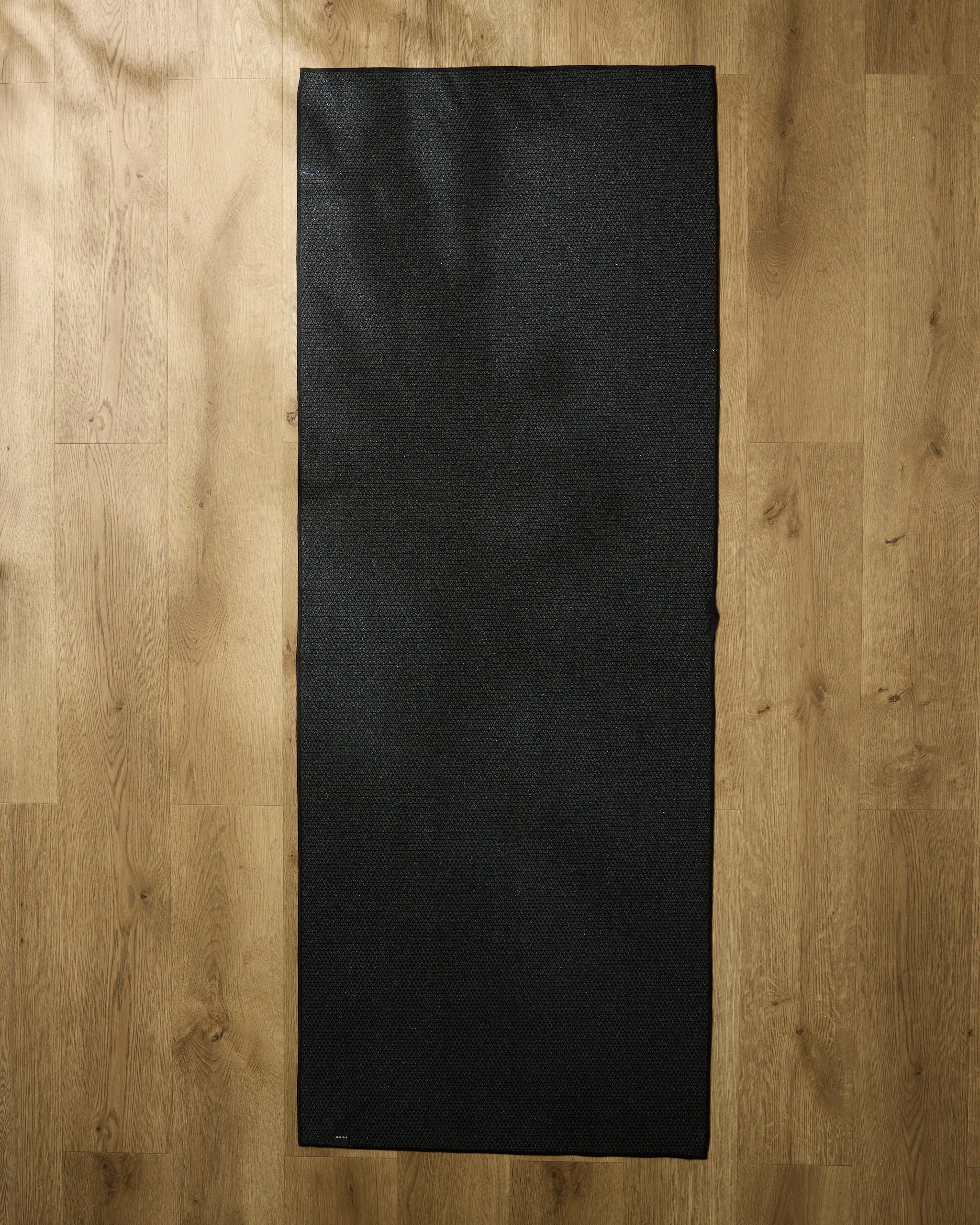The Geometry Airy Flow rectangular black yoga mat with a recycled towel is laid flat on a light wooden floor, seen from above.