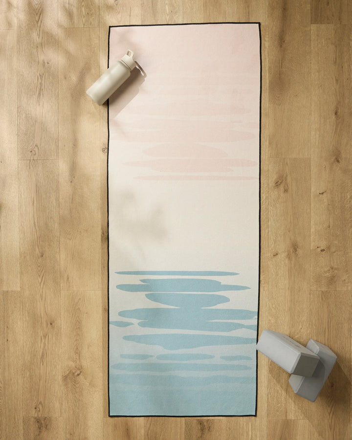The GEOMETRY Balance yoga mat with a light pink to light blue gradient rests on a wooden floor, accompanied by a non-slip yoga towel, water bottle, and two yoga blocks in the mat’s corners.