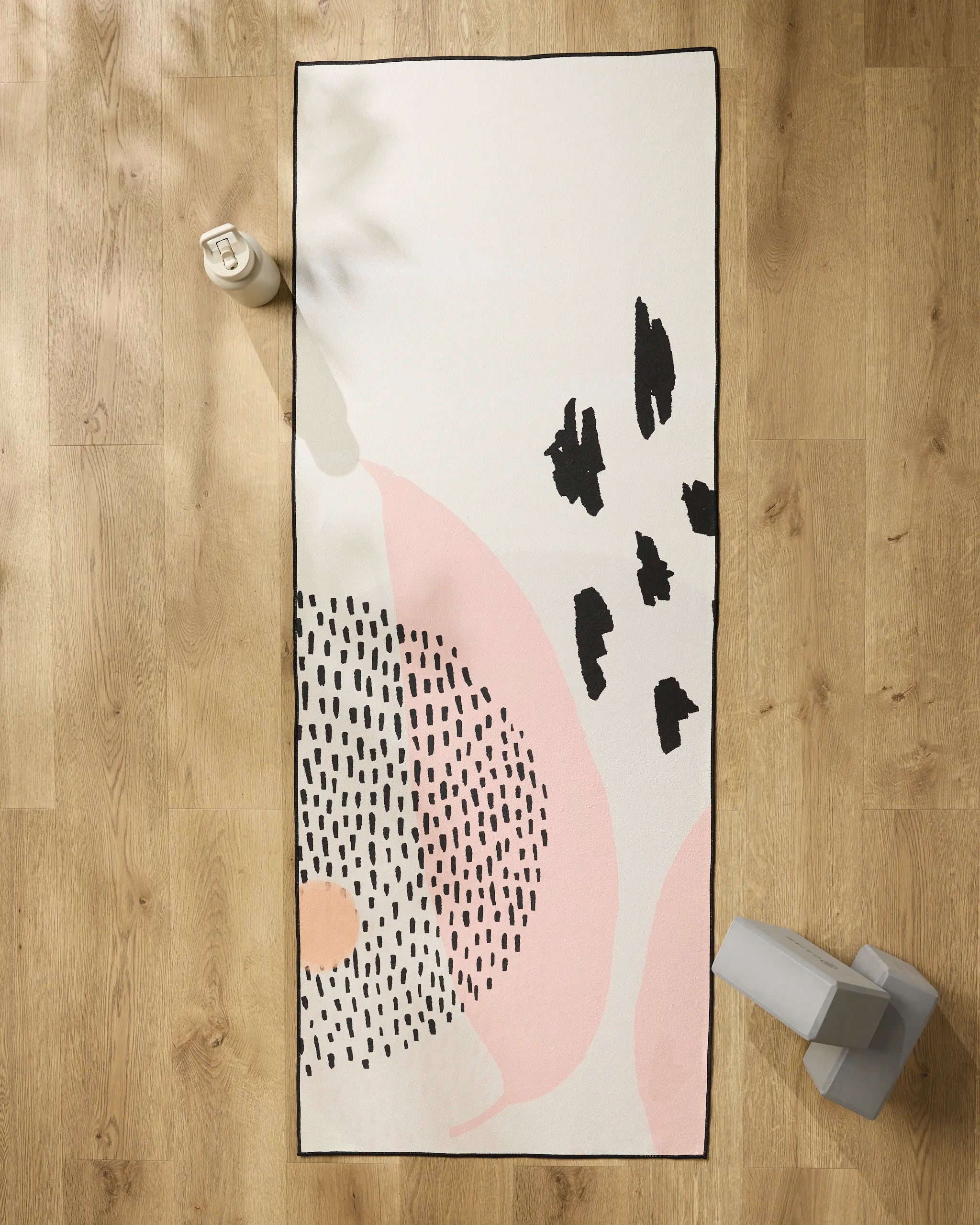 The GEOMETRY Bare rectangular recycled yoga towel, featuring abstract black and pink patterns, is laid on a wooden floor with a white water bottle at the top left and gray weights at the bottom right.