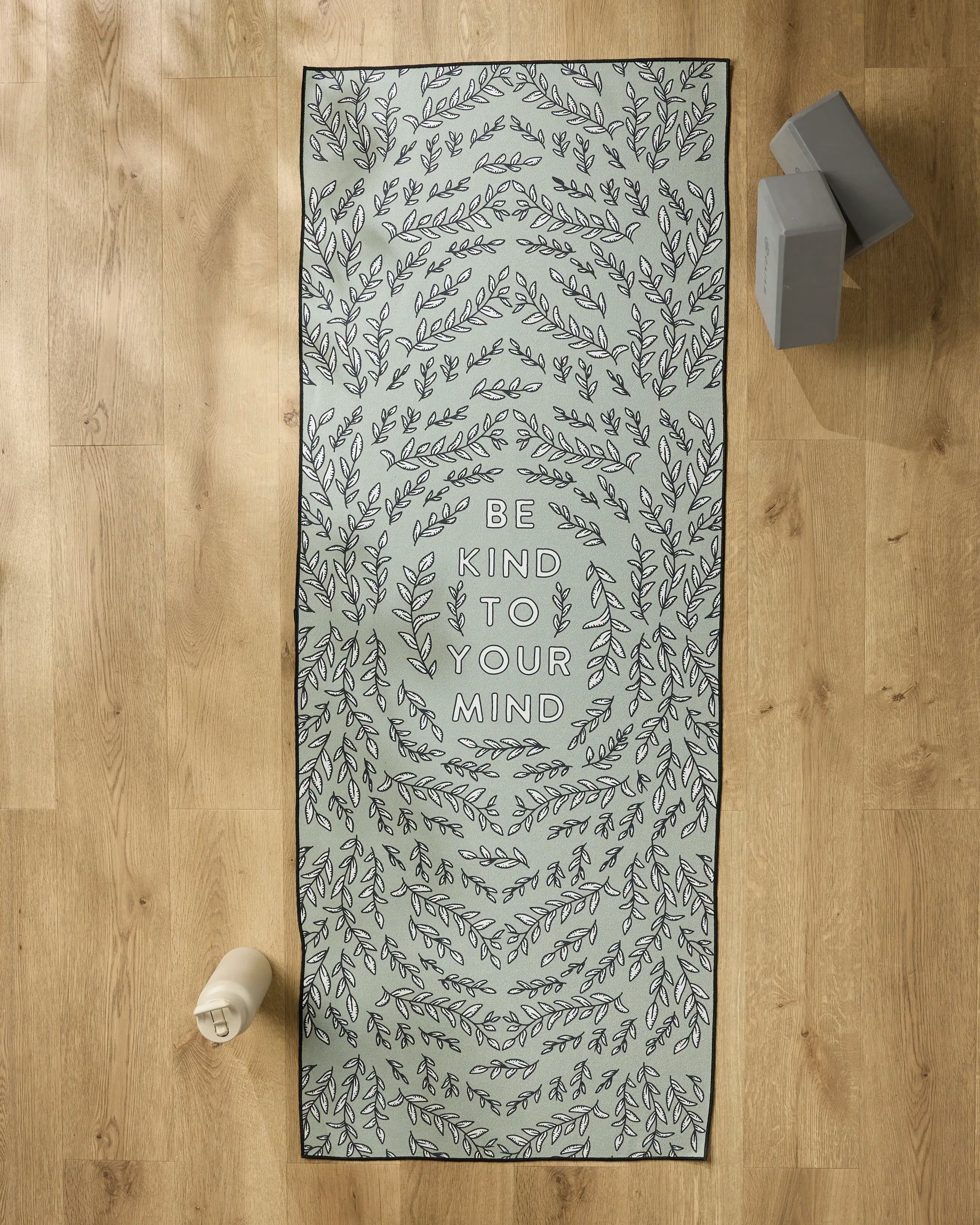The Graphics and Grain "Be Kind To Your Mind" yoga mat with leafy patterns rests on a wooden floor, paired with a slip-resistant yoga towel, water bottle, and two stacked yoga blocks.