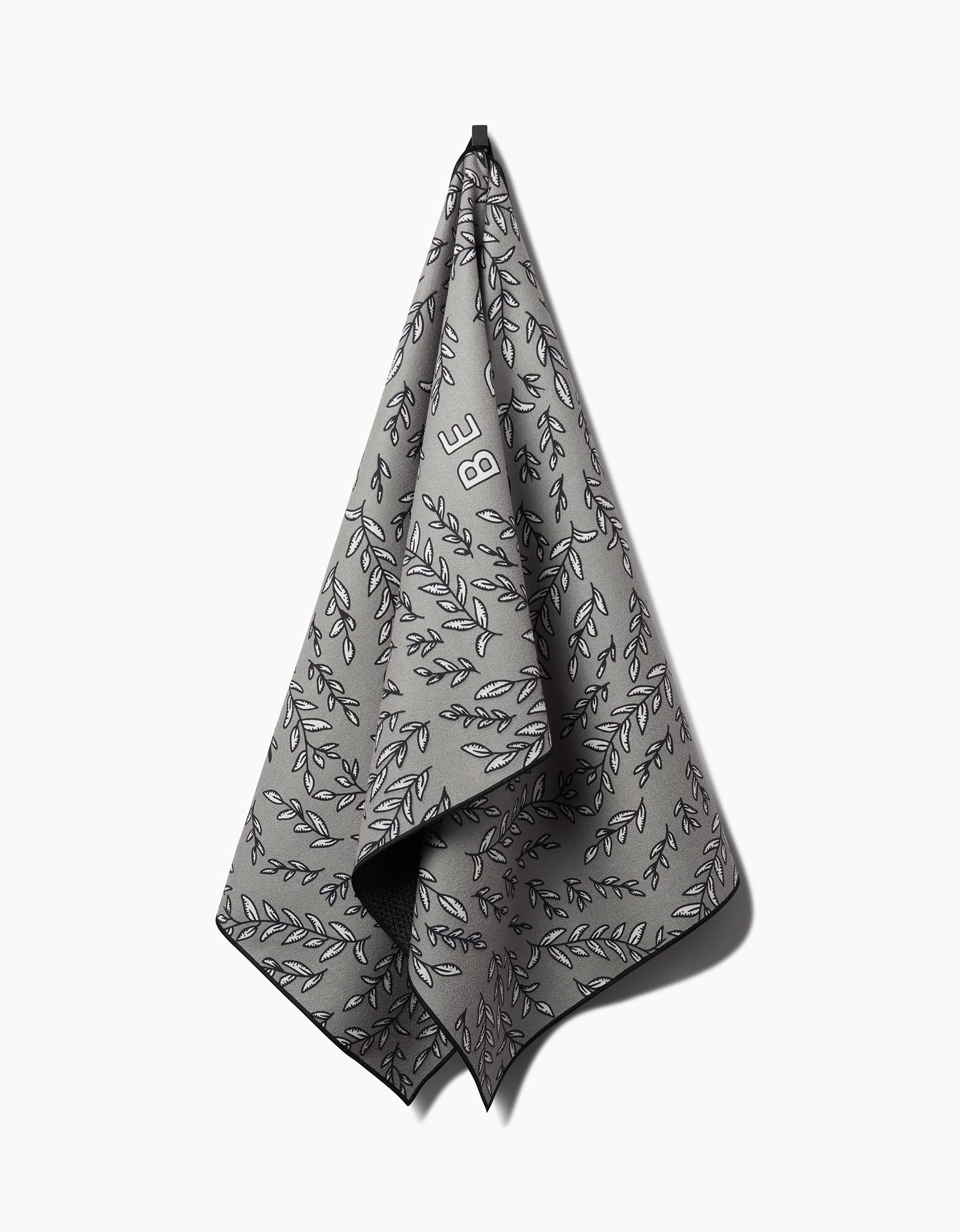 The Be Kind To Your Mind yoga towel by Graphics and Grain, crafted from recycled materials, features a black leafy pattern with the word BE in white on gray fabric. It hangs on a hook against a white background.