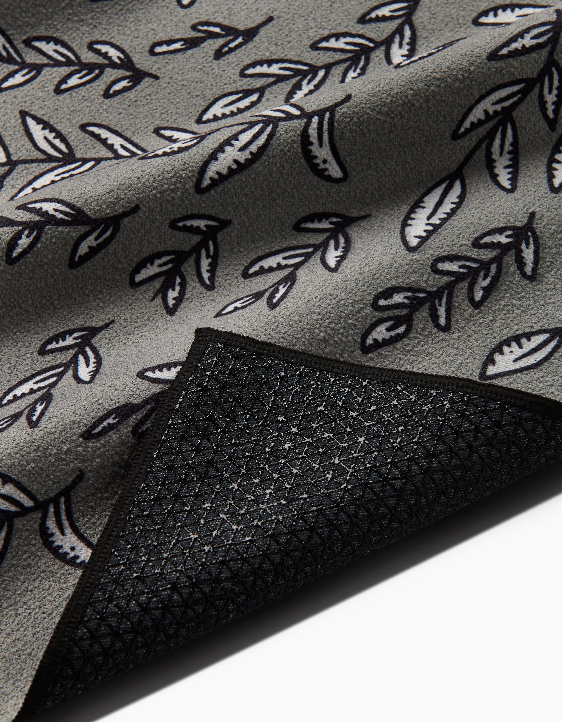A close-up of the Be Kind To Your Mind mat by Graphics and Grain, featuring a gray fabric with a black and white leaf pattern made from recycled materials. The flipped corner reveals its textured, slip-resistant black underside.