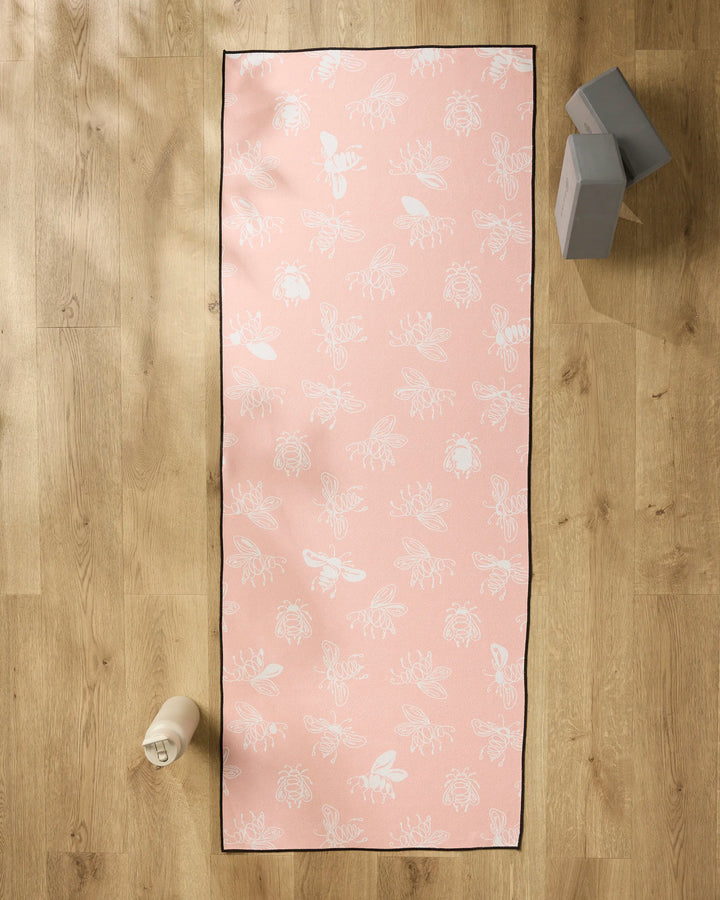 The Sara Mari Beelieve recycled pink yoga mat towel with white bee and flower patterns is on a wooden floor, next to a white water bottle and two gray blocks stacked at the top right.