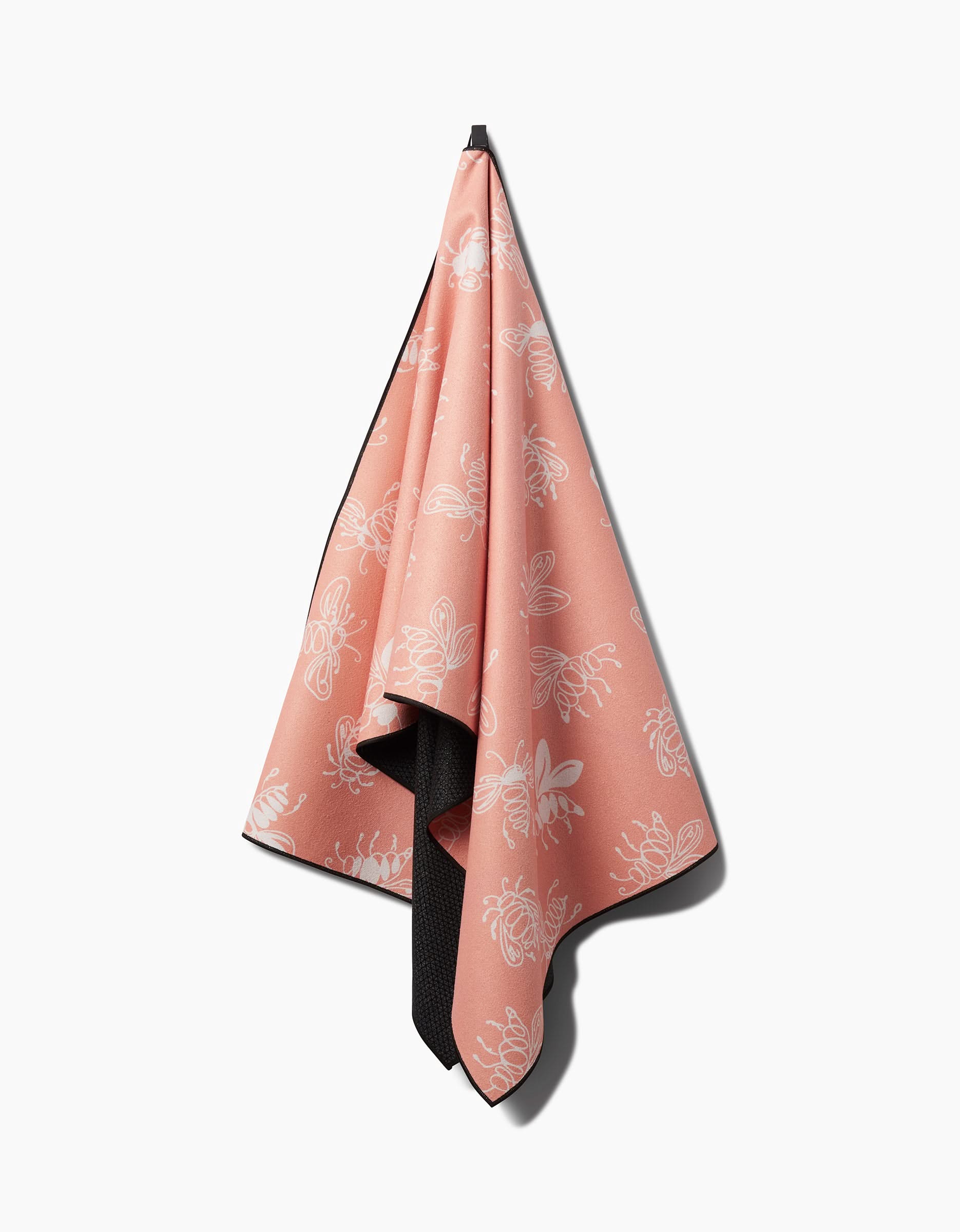 The Sara Mari Beelieve yoga towel features soft pink fabric with white bee and floral patterns, a dark slip-resistant underside, and hangs from a hook against a white background—perfect for steady and stylish practice.
