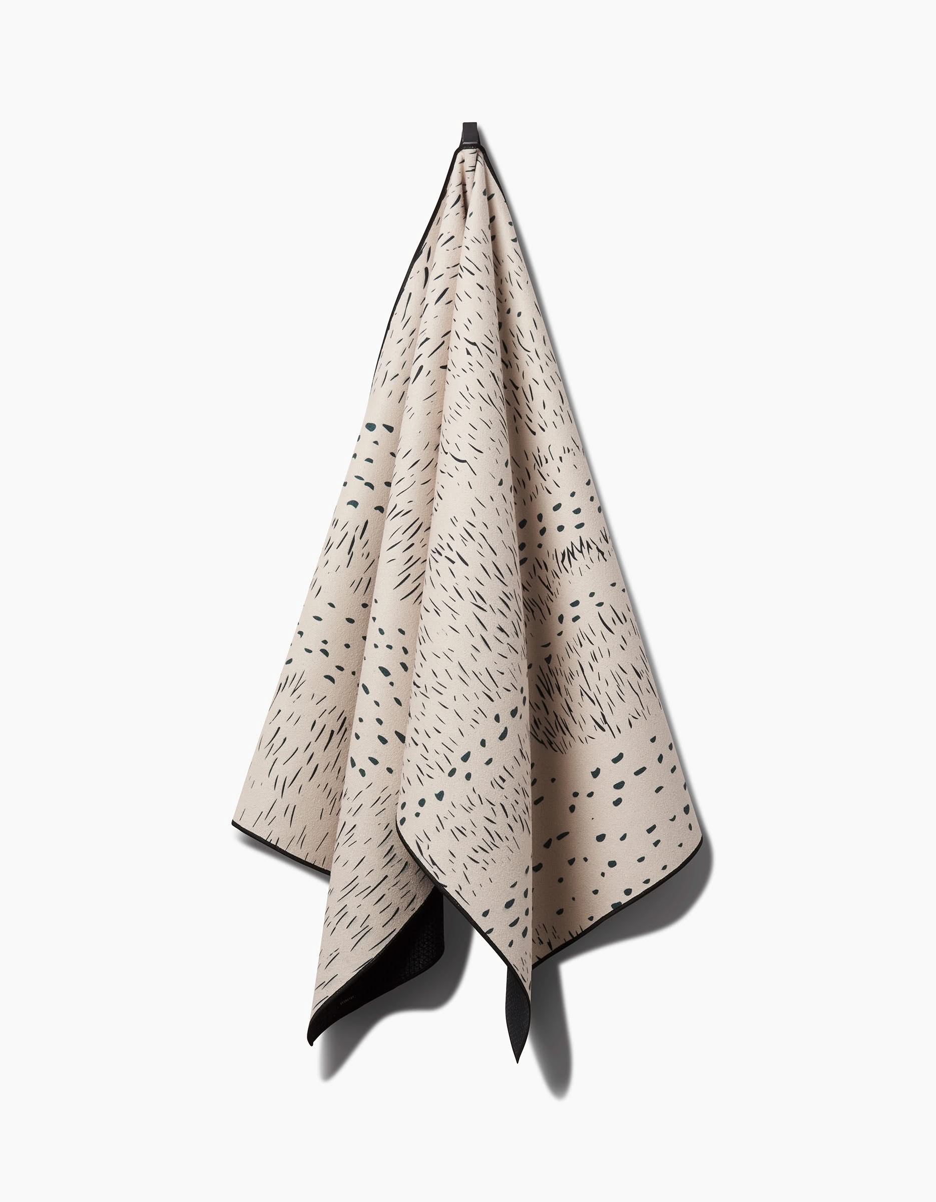 The GEOMETRY Bend yoga towel, made from recycled fabric in beige with black irregular dash and dot patterns and black edging, hangs neatly on a hook against a white background, its soft drape casting a gentle shadow.