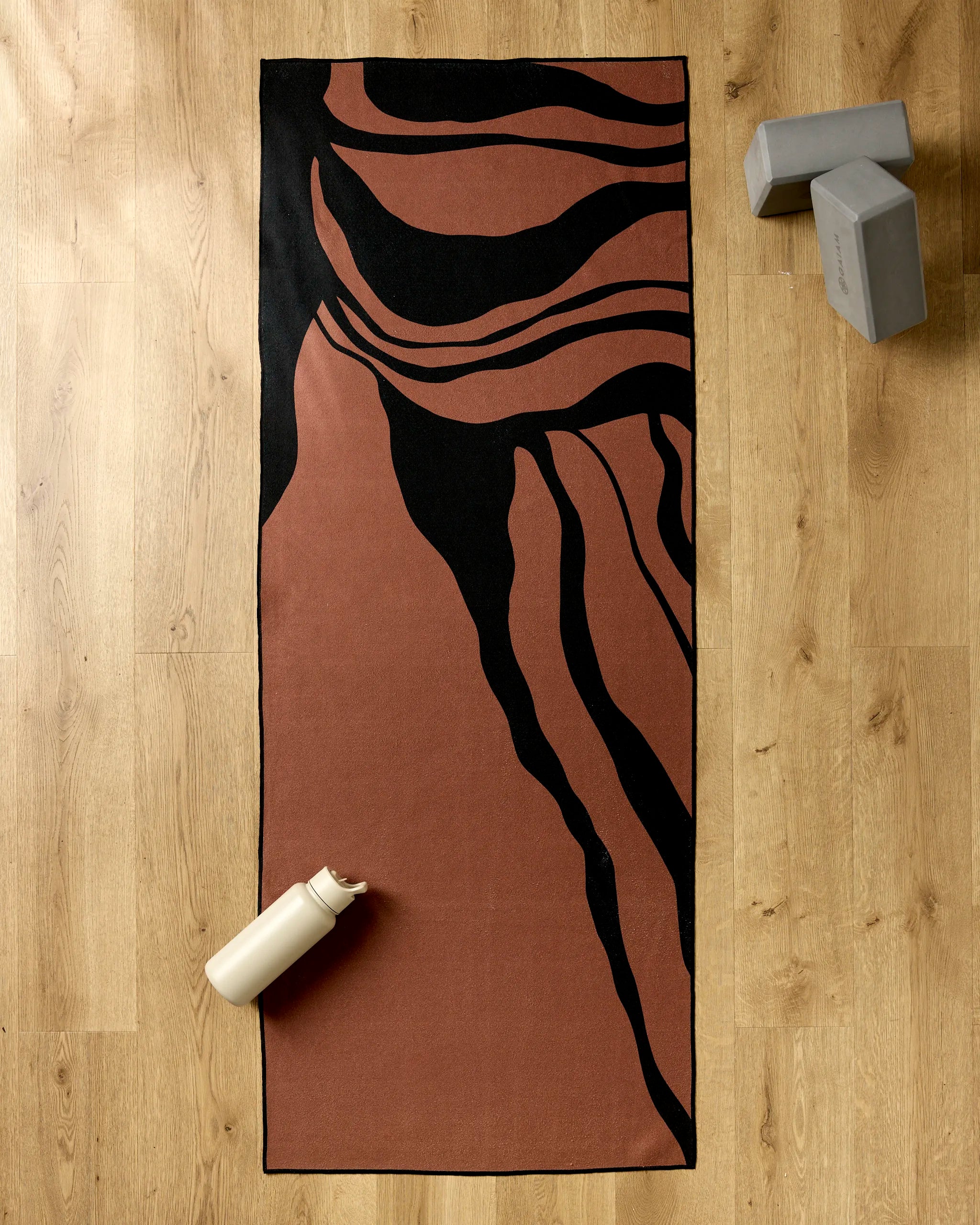 The Cole Strem Black Swirl yoga mat is on a wooden floor with a cream water bottle to the left, two gray yoga blocks stacked at the top right, and a non-slip yoga towel neatly folded nearby.