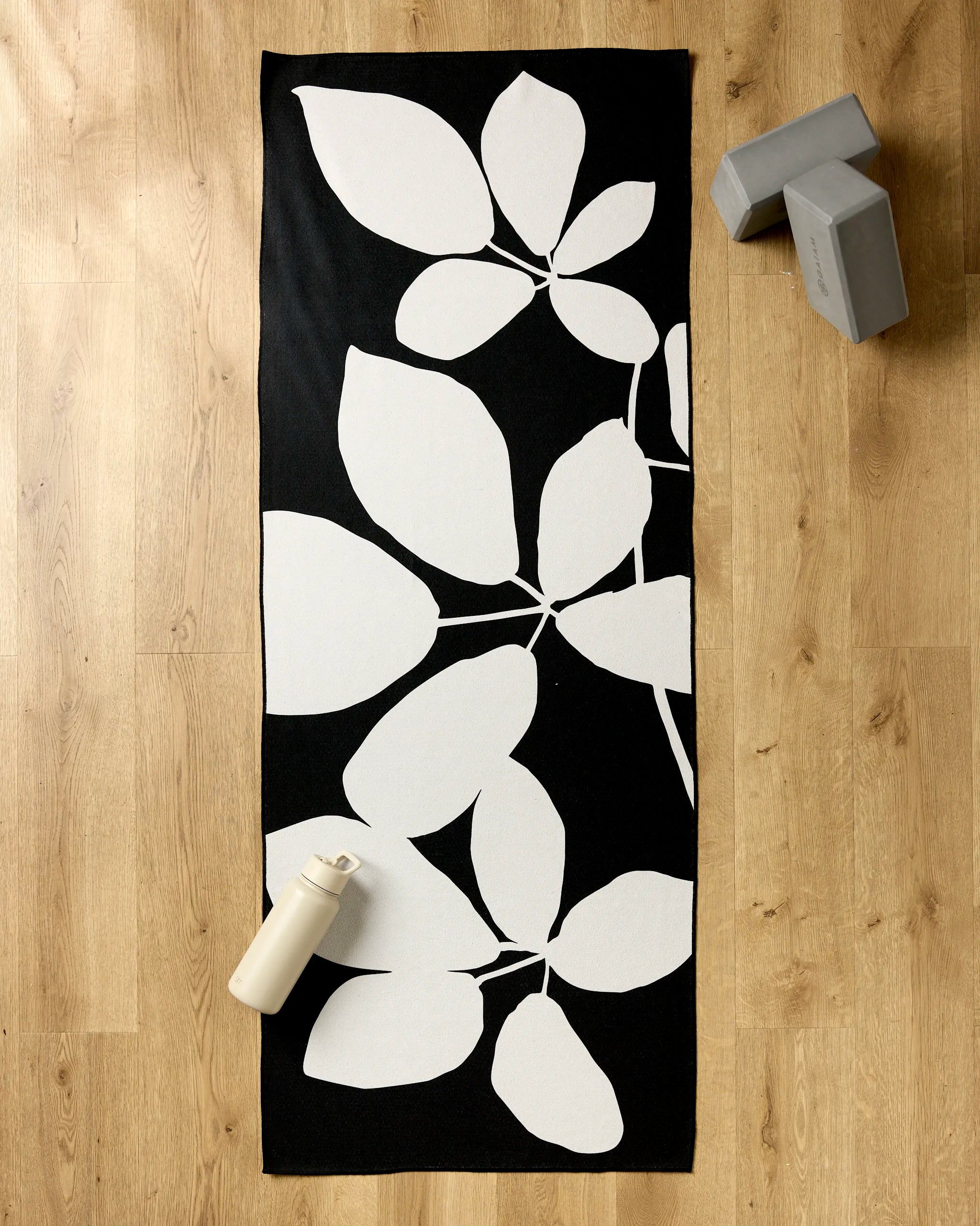 The PrintedPlant "Blackberry" yoga mat with a white leaf pattern rests on a light wooden floor, accompanied by a cream water bottle, two gray yoga blocks, and a non-slip towel nearby.
