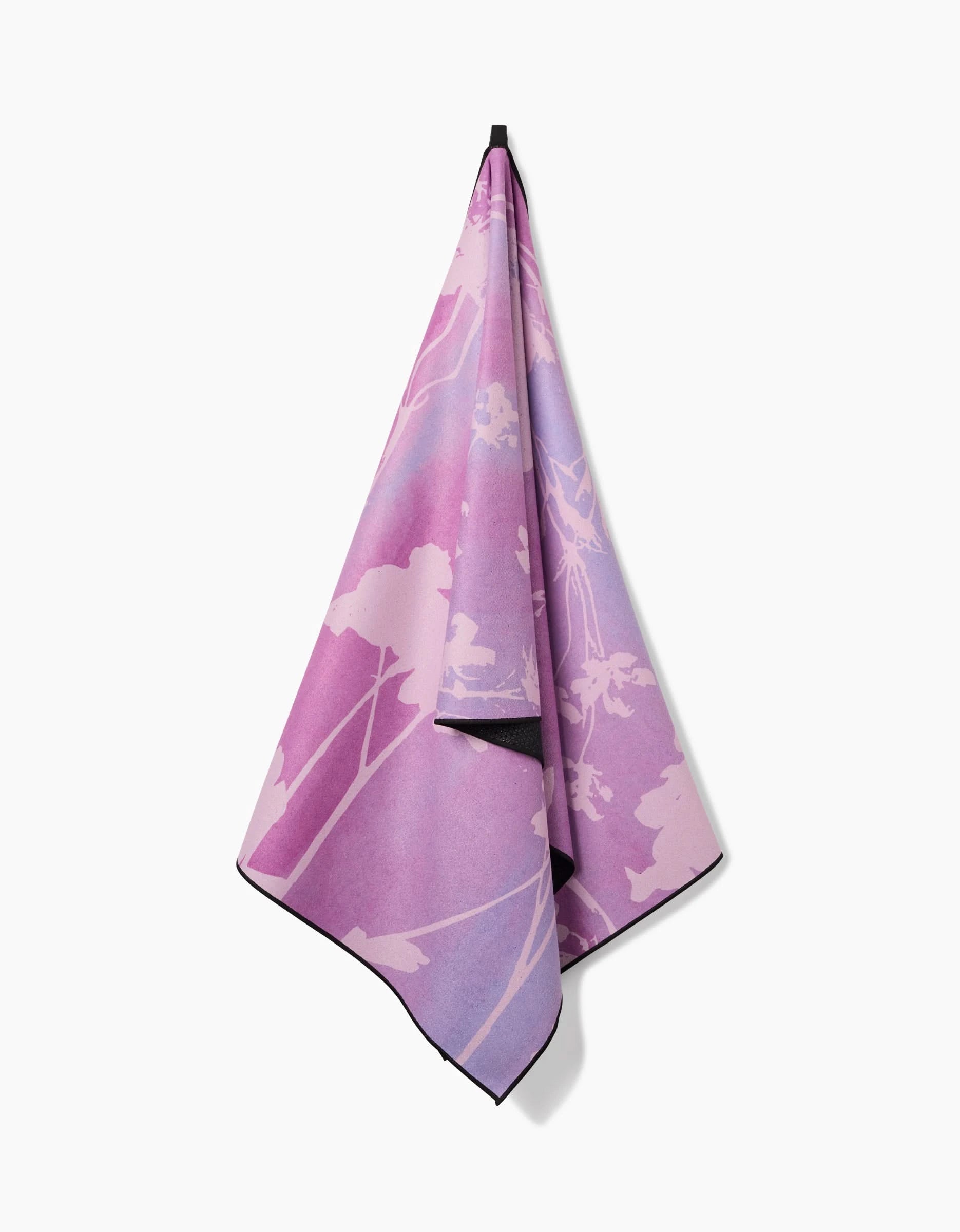 The GEOMETRY Bloom Blend yoga towel, featuring purple and pink hues with a subtle floral design and made from recycled materials, hangs on a hook against a plain white background.