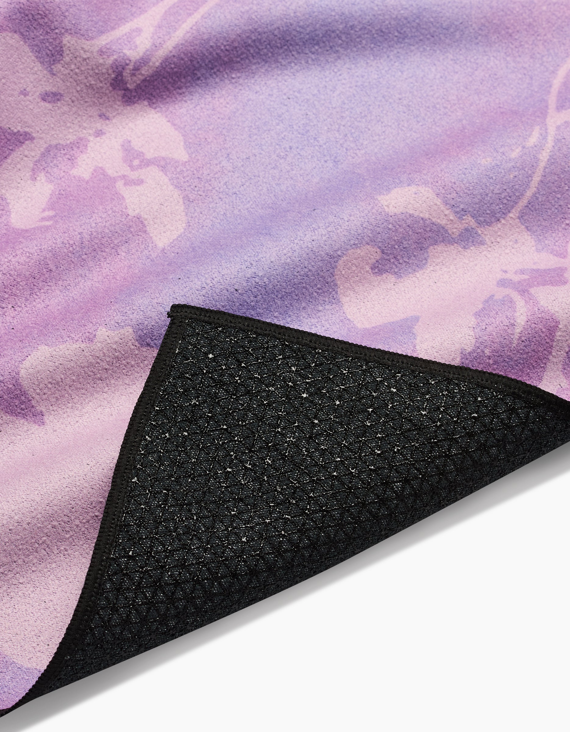 A close-up of the GEOMETRY Bloom Blend yoga mat, featuring a purple and pink floral pattern, crafted from recycled materials with a black textured non-slip backing; one corner is slightly lifted to show the underside.