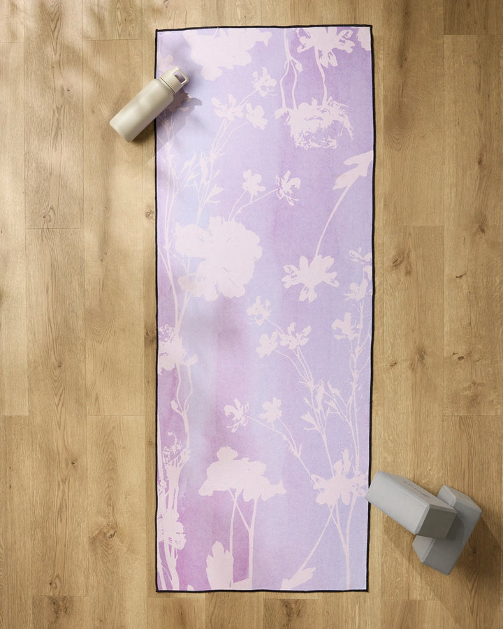 The GEOMETRY Bloom Blend yoga mat with white floral patterns is on a wooden floor, topped with a silicone grip yoga towel. A water bottle sits at the top left and gray yoga blocks are at the bottom right of the mat.