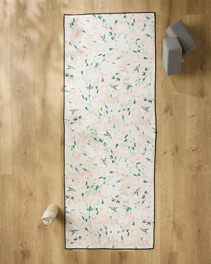 The Blossoms yoga mat by Anee Shah, featuring green, pink, and white florals, rests on a wooden floor with a white water bottle, two gray blocks stacked nearby, and a soft non-slip yoga towel for extra comfort.
