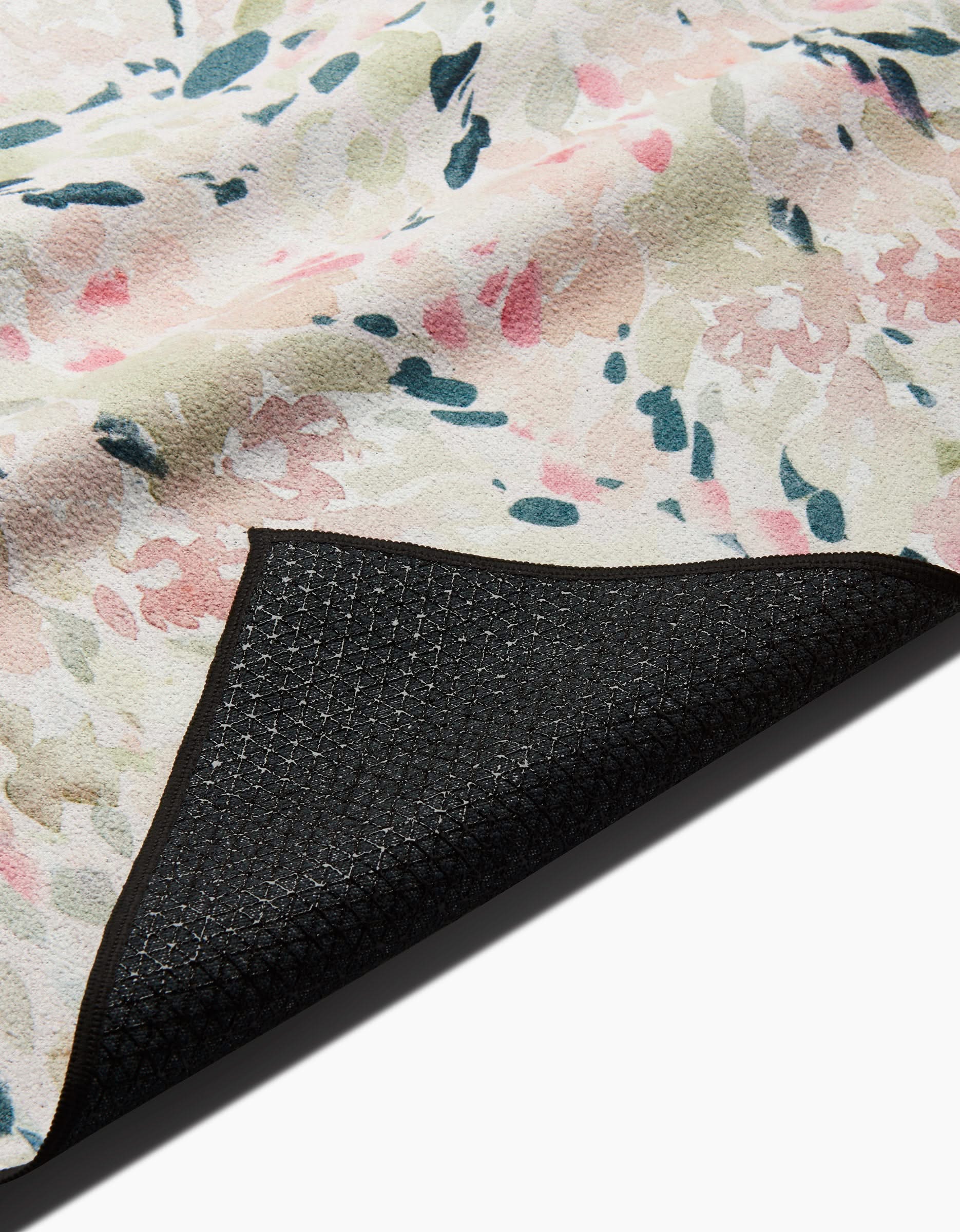 A close-up of the Blossoms yoga towel by Anee Shah, featuring a floral pattern in pale pink, green, and blue. One corner is flipped to show its textured black silicone grip backing for secure yoga practice.