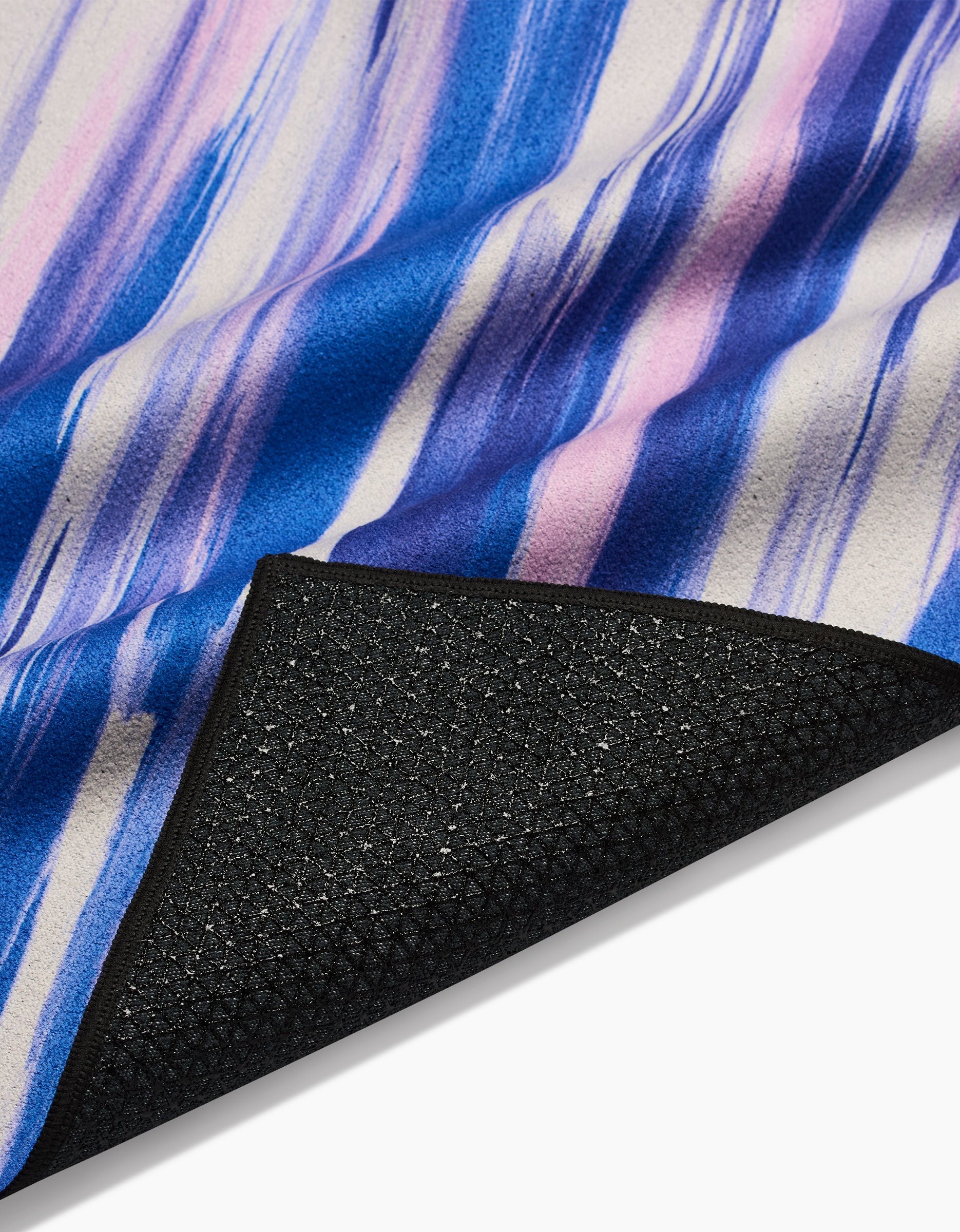 A close-up of GEOMETRYs Blue Blast yoga towel, featuring blue, purple, and white watercolor stripes. The flipped corner reveals a textured black underside made from recycled materials.