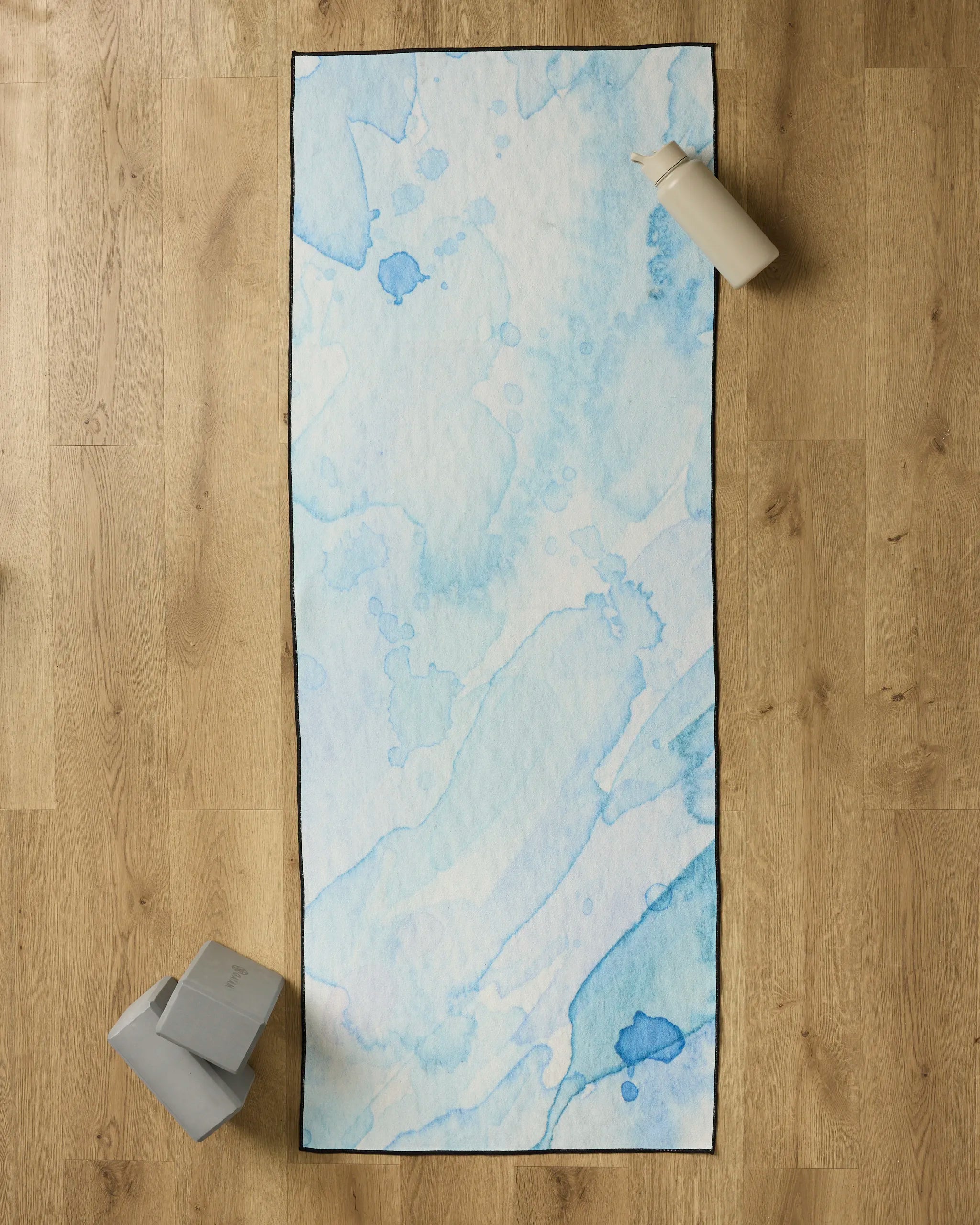 The GEOMETRY Boundless Blue yoga towel, featuring a blue and white watercolor pattern, is spread on a light wooden floor. A white water bottle sits at the top right and two gray yoga blocks rest at the bottom left.