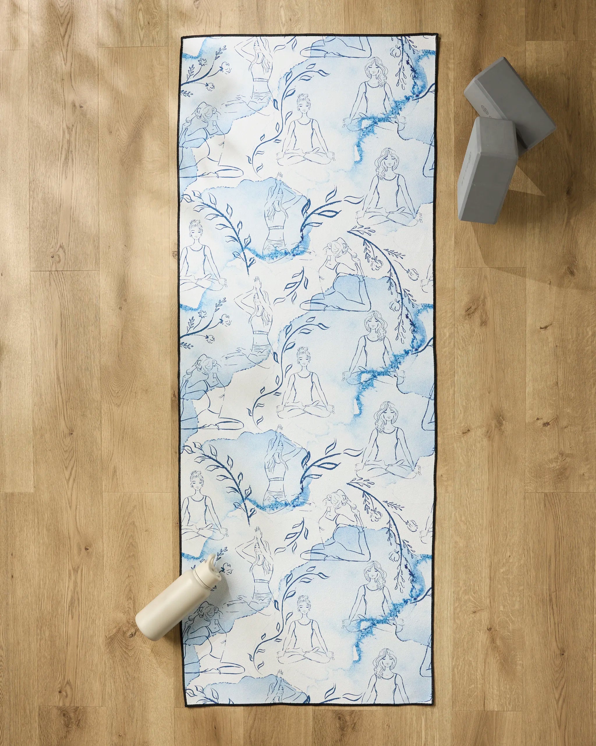 The "Breathe In Breathe Out" yoga mat by Elena Fay, featuring blue sketches of women in yoga poses and leafy branches, rests on a wooden floor beside a white water bottle, two gray blocks, and an eco-friendly yoga towel.