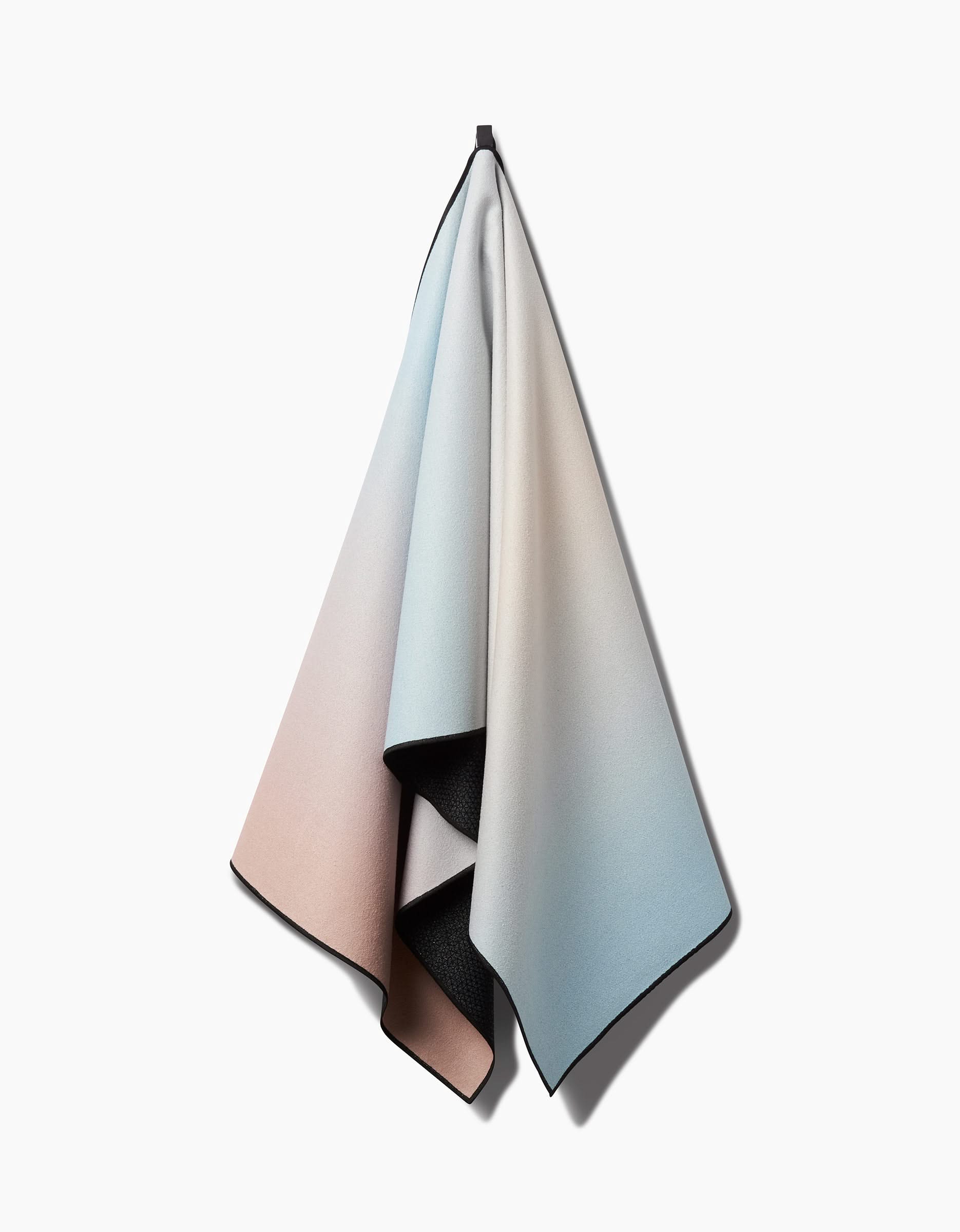 The GEOMETRY Breathe yoga towel, featuring a pink-to-cream-to-blue gradient and slip-resistant softness, hangs from a hook against a plain white background.