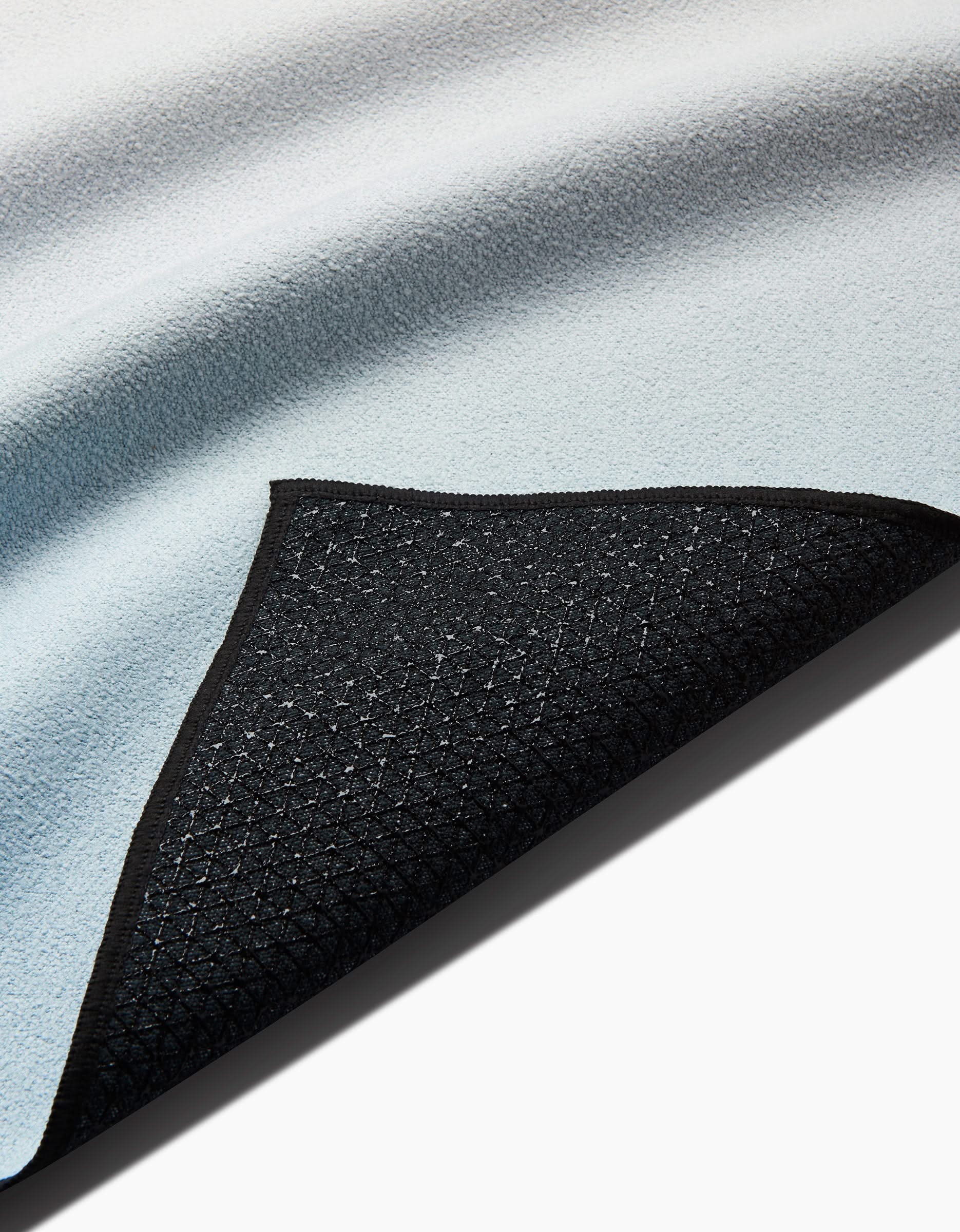 A close-up of GEOMETRY’s Breathe yoga towel in light blue, featuring a corner flipped to show its black, slip-resistant diamond-patterned backing. This recycled towel rests on a white surface.