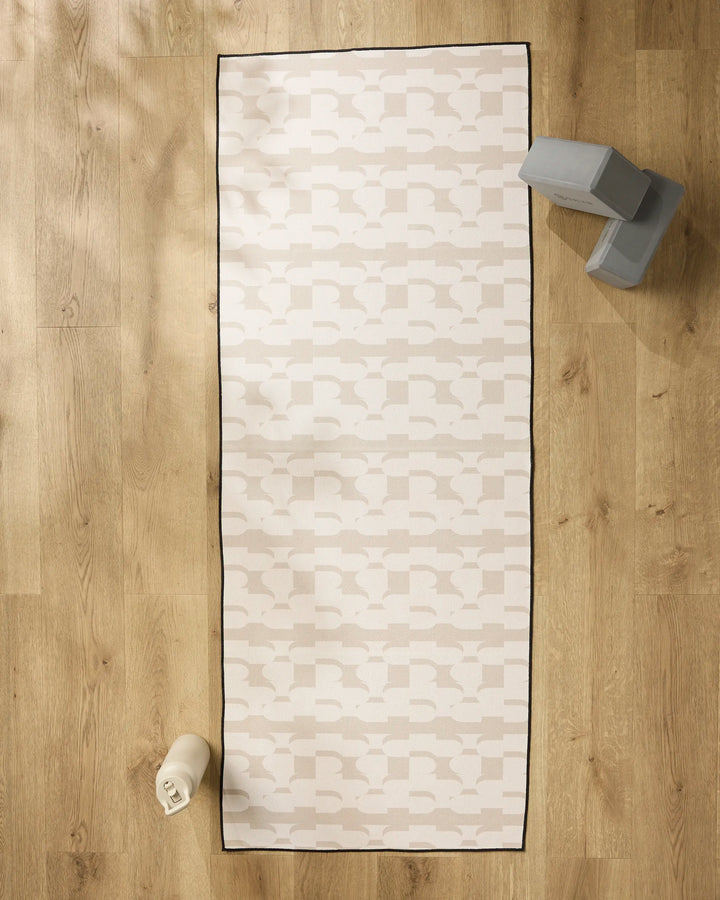 A cream Buttons yoga mat by Geometry with a subtle geometric pattern rests on a wooden floor, accompanied by a gray yoga block and a rolled recycled yoga towel—creating an inviting space for your practice.