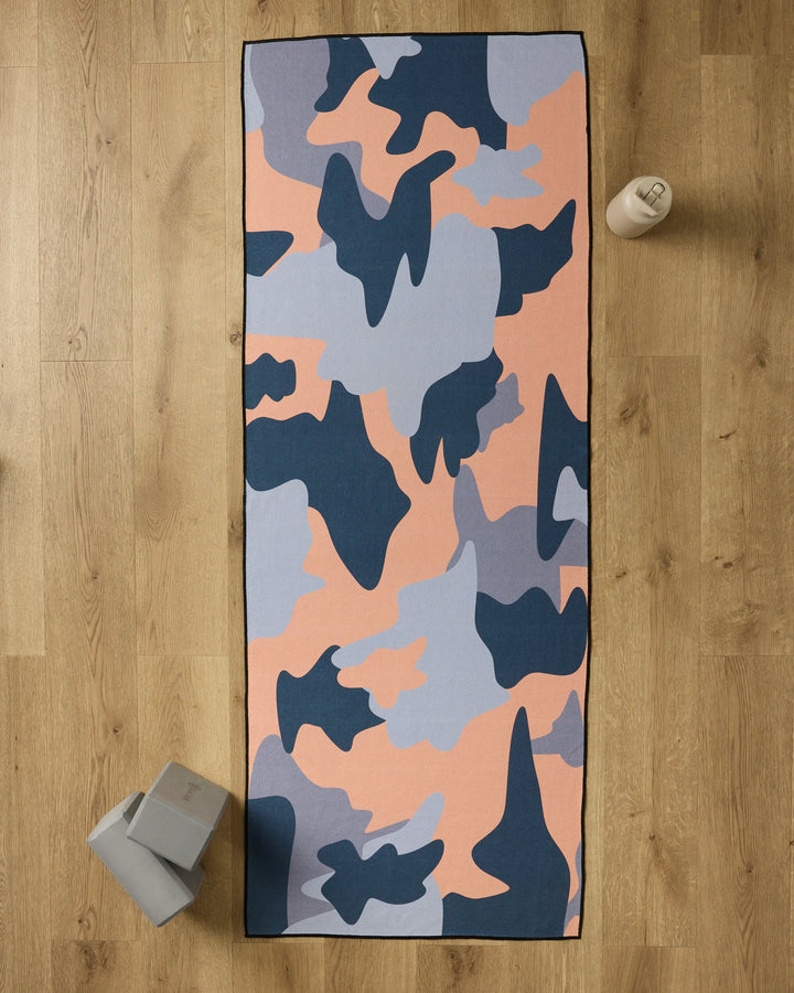 The GEOMETRY Camo yoga towel, featuring a pink, blue, and navy camouflage pattern, is spread on a wooden floor with a water bottle at the top right and two gray yoga blocks at the bottom left.