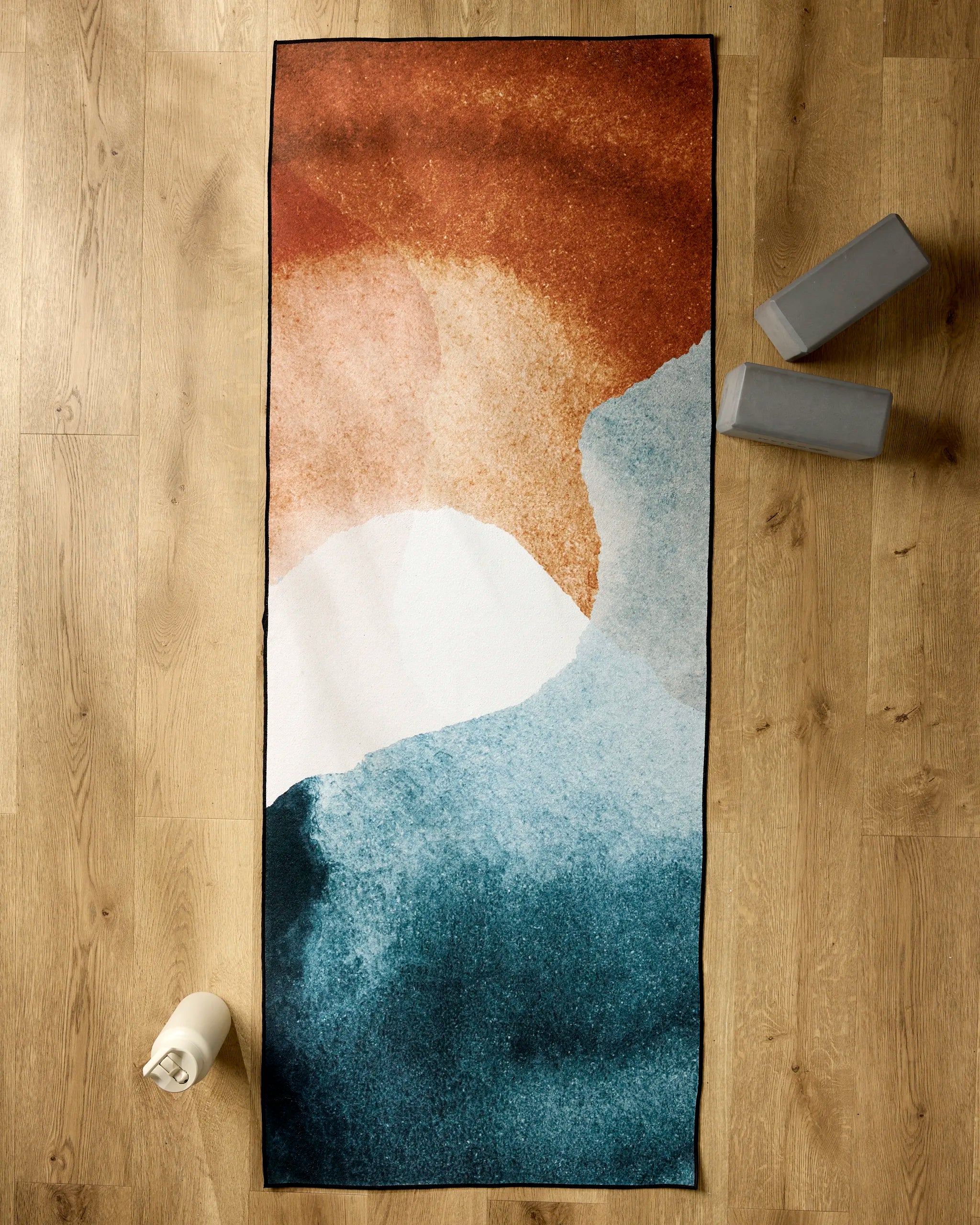 GEOMETRY’s Centered yoga towel, featuring an abstract blue, white, and rust design, rests on a wooden floor beside two gray yoga blocks and a white water bottle.