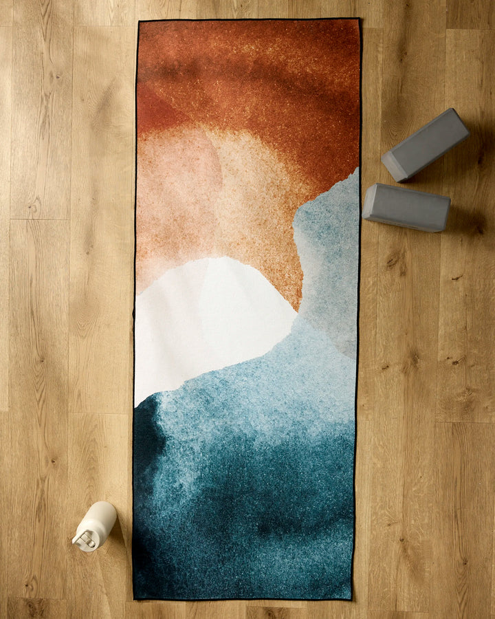 GEOMETRY’s Centered yoga towel, featuring an abstract blue, white, and rust design, rests on a wooden floor beside two gray yoga blocks and a white water bottle.