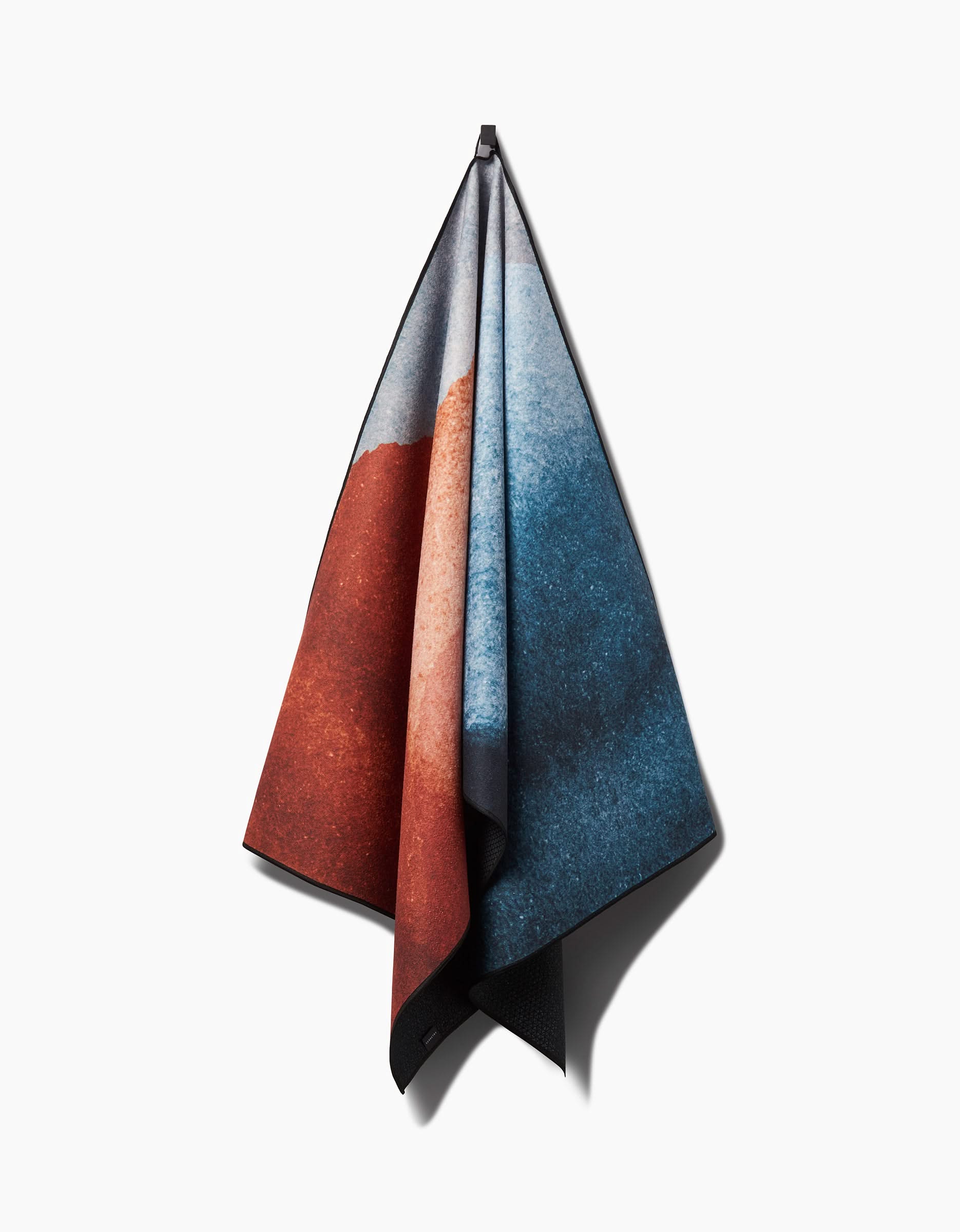 The GEOMETRY Centered yoga towel, featuring an abstract design in red, blue, and gray tones, hangs from a single hook against a plain white background.