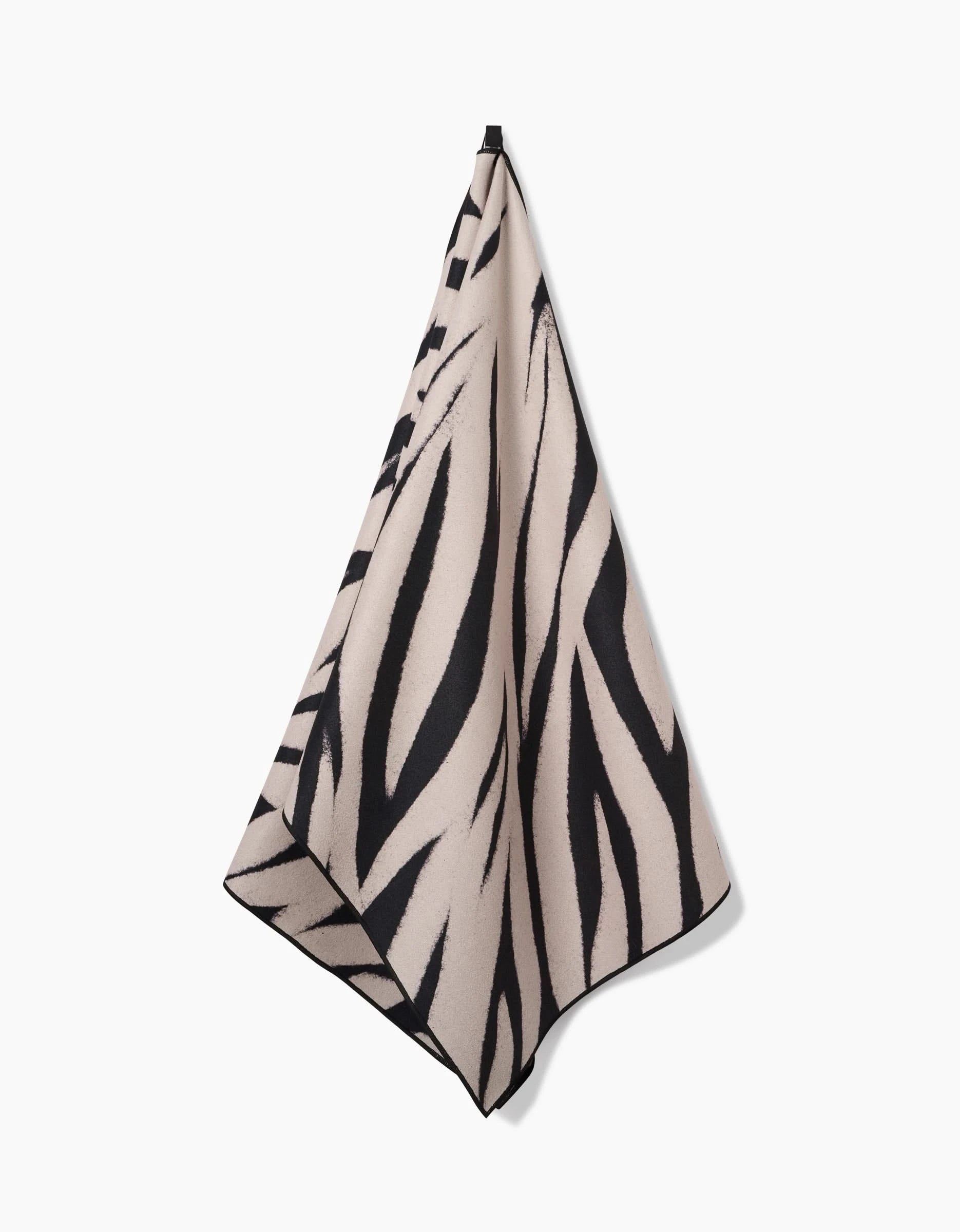 The GEOMETRY Chase yoga towel, featuring bold black zebra stripes on beige and made from recycled materials, hangs from a single point against a plain white background.