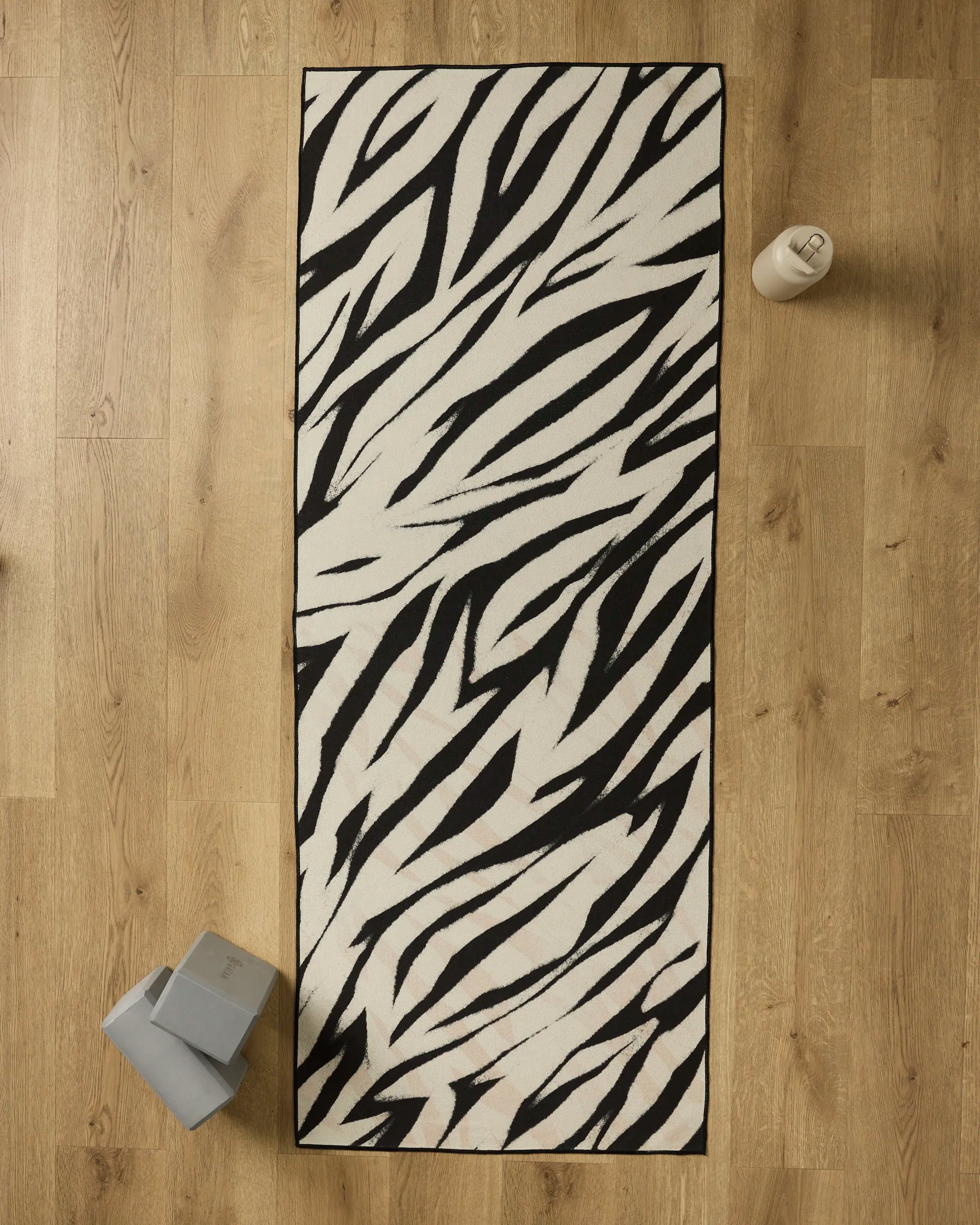 The GEOMETRY Chase yoga mat with a black and white zebra stripe pattern lies on a wooden floor, topped with a non-slip towel. Gray yoga blocks and a beige water bottle are placed nearby.