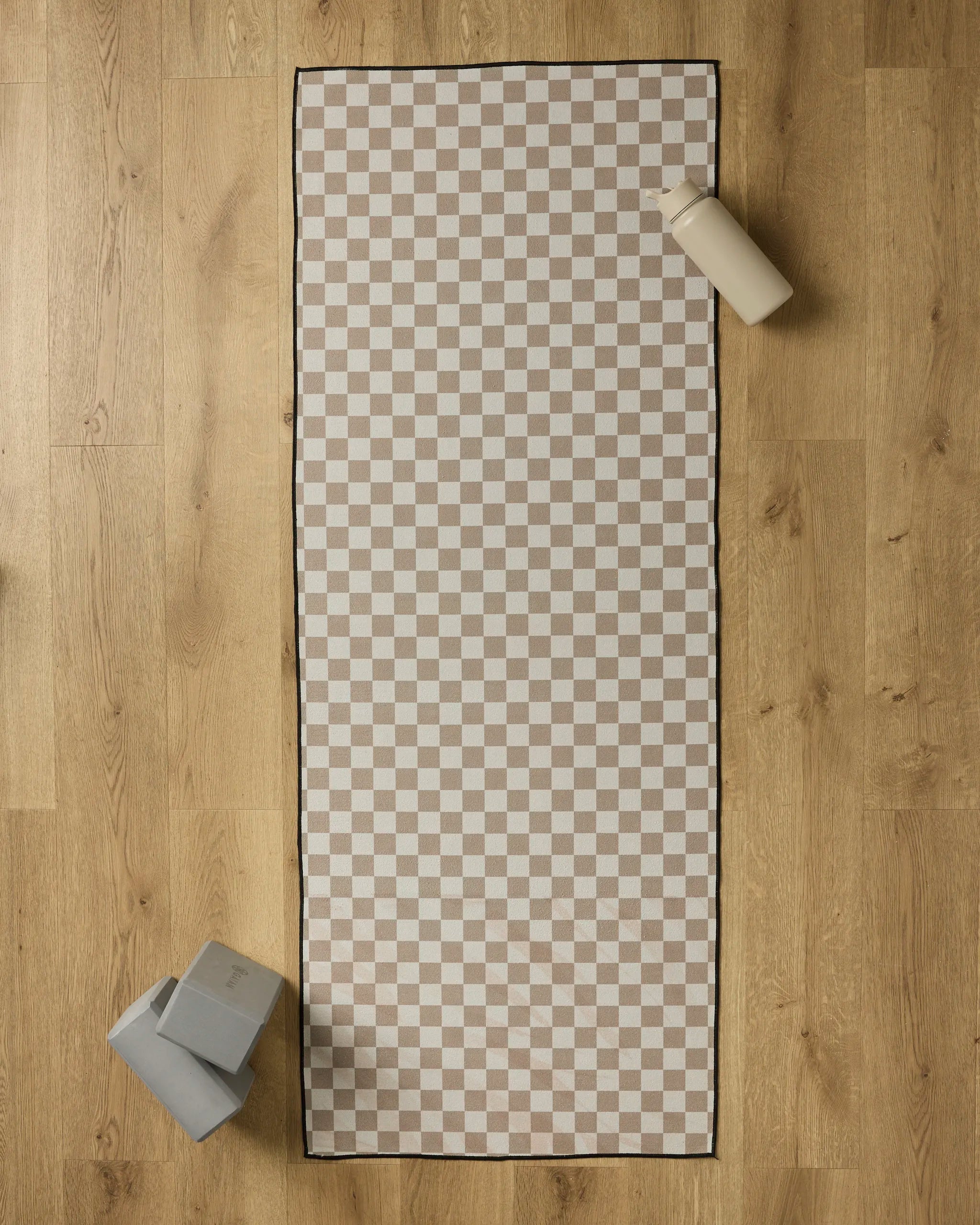 The Chic Checker yoga mat towel by Geometry, featuring a beige and white checkered design, is laid on a wooden floor with a water bottle in the top right corner and two stacked yoga blocks in the bottom left.