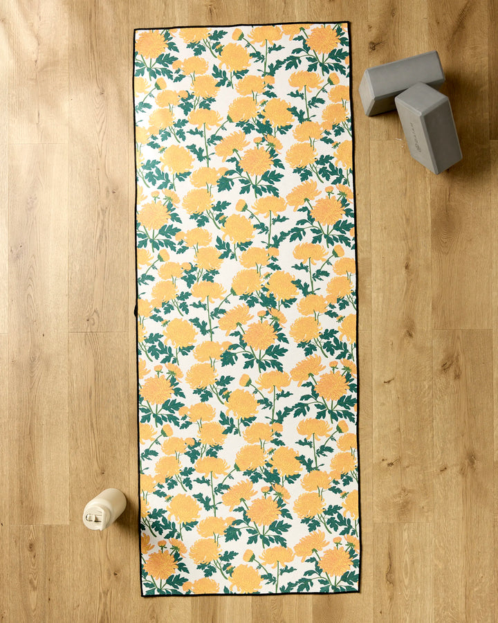 The Chrysanthemum yoga mat from Vaishali Design Studio, featuring yellow and green floral patterns, lies on a wooden floor with gray yoga blocks, a light water bottle, and a non-slip towel beside it for added comfort.