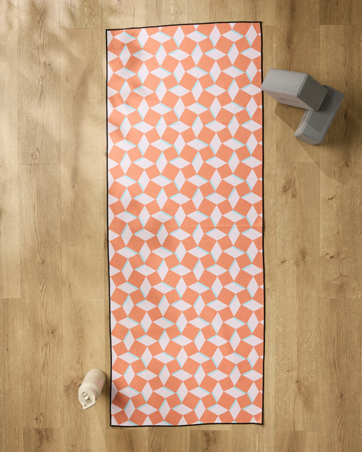 The Geometry Circus Stand Pink Orange yoga mat is laid out on a wooden floor, with a non-slip yoga towel at one end, a water bottle, and two stacked gray yoga blocks beside it.