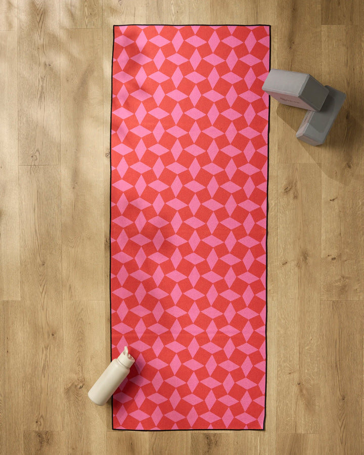 The Geometry Circus Stand Pink Red yoga mat lies on a wooden floor, with two gray yoga blocks, a white water bottle, and a non-slip yoga towel placed nearby.