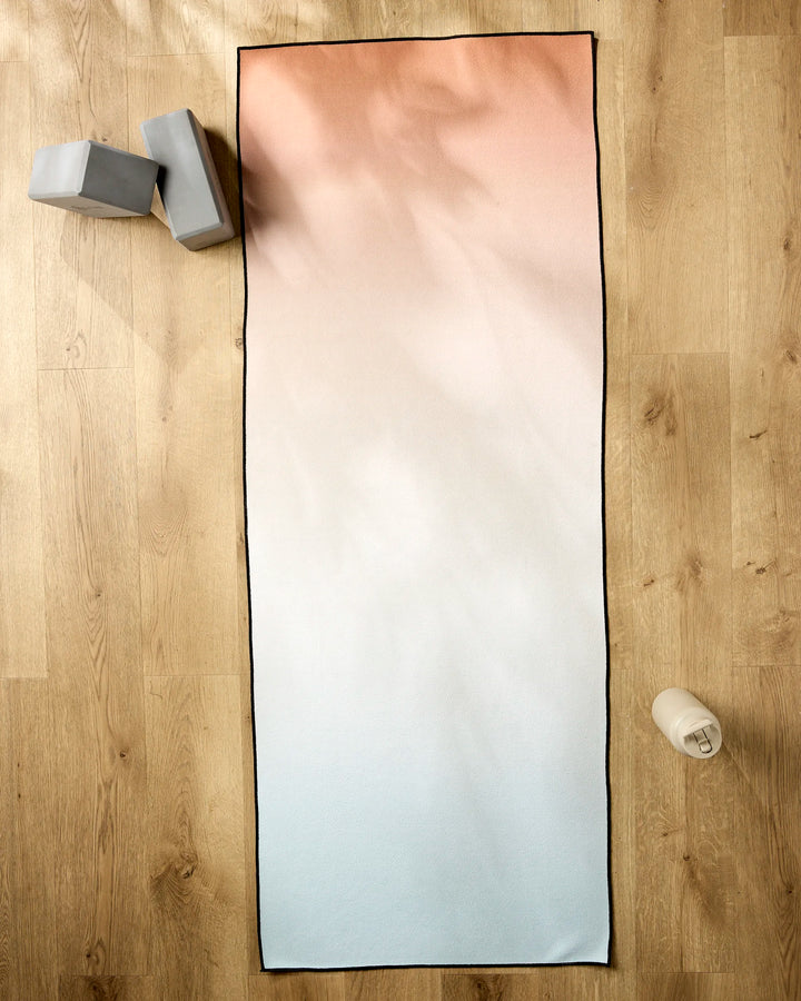 The GEOMETRY Core yoga mat, featuring a gradient from light pink to light blue, rests on a wooden floor alongside two gray foam blocks, a white water bottle, and a non-slip yoga towel.