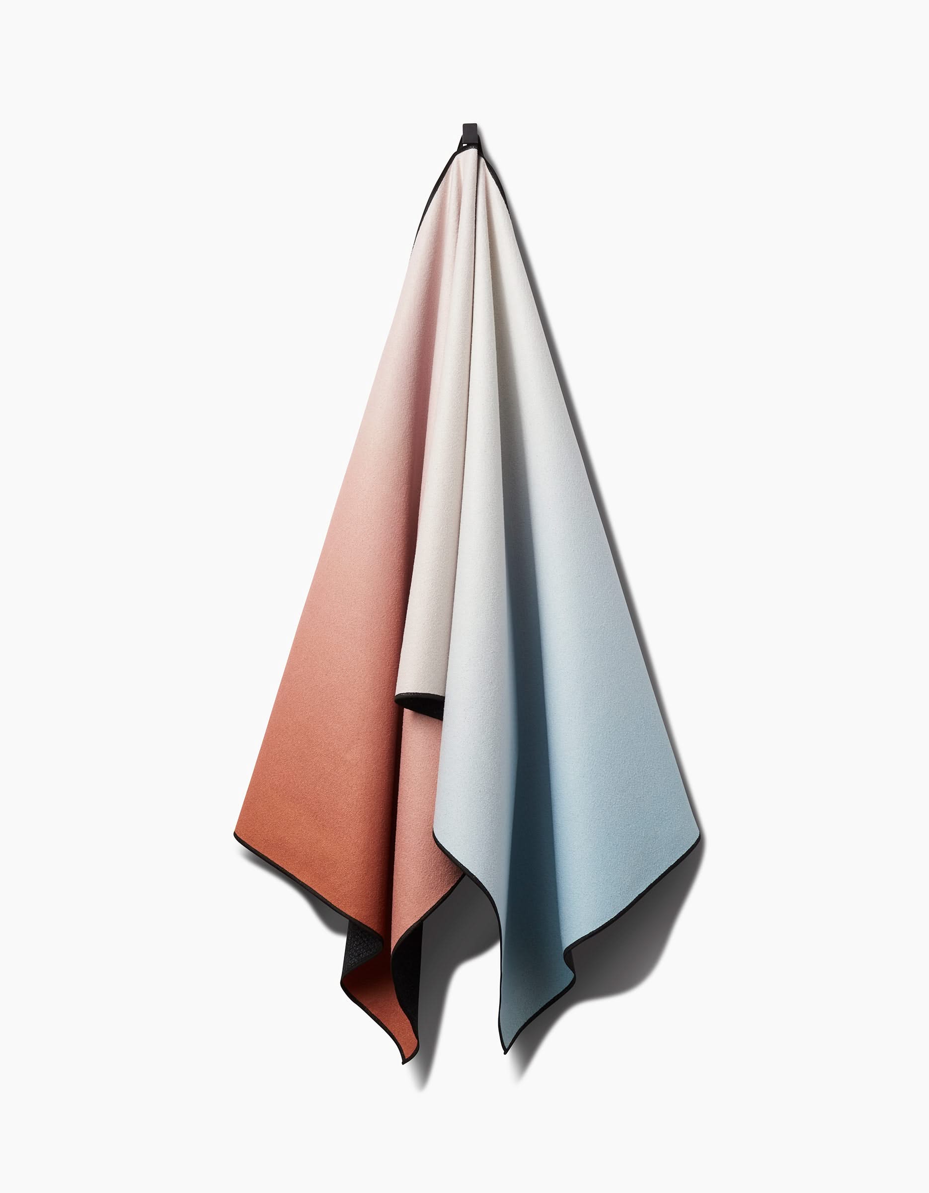 The GEOMETRY Core yoga towel, featuring a gradient from rust orange to off-white to pale blue, hangs on a white wall and casts a soft shadow.