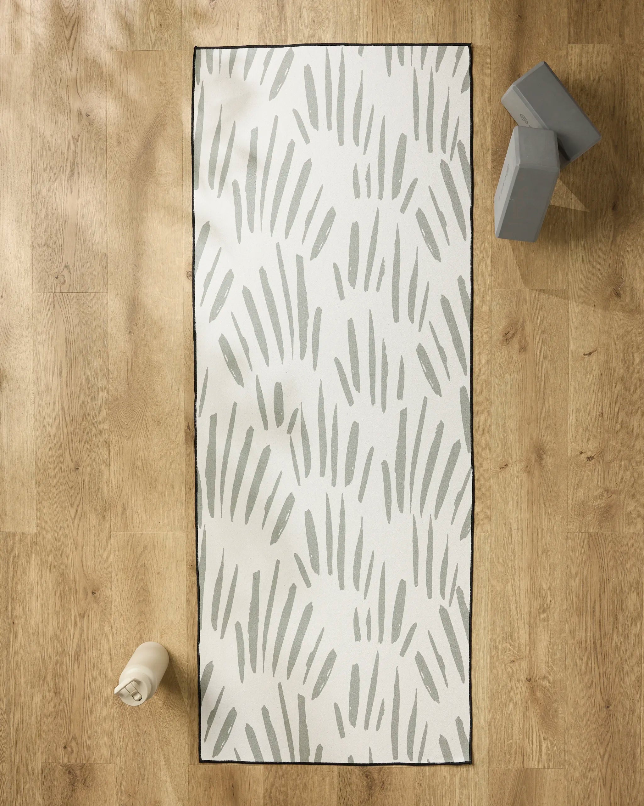 The Country Fields yoga mat by Hufton Studio, featuring gray abstract brushstrokes, rests on a light wood floor with a non-slip towel, water bottle, and two gray yoga blocks nearby for a serene workout space.