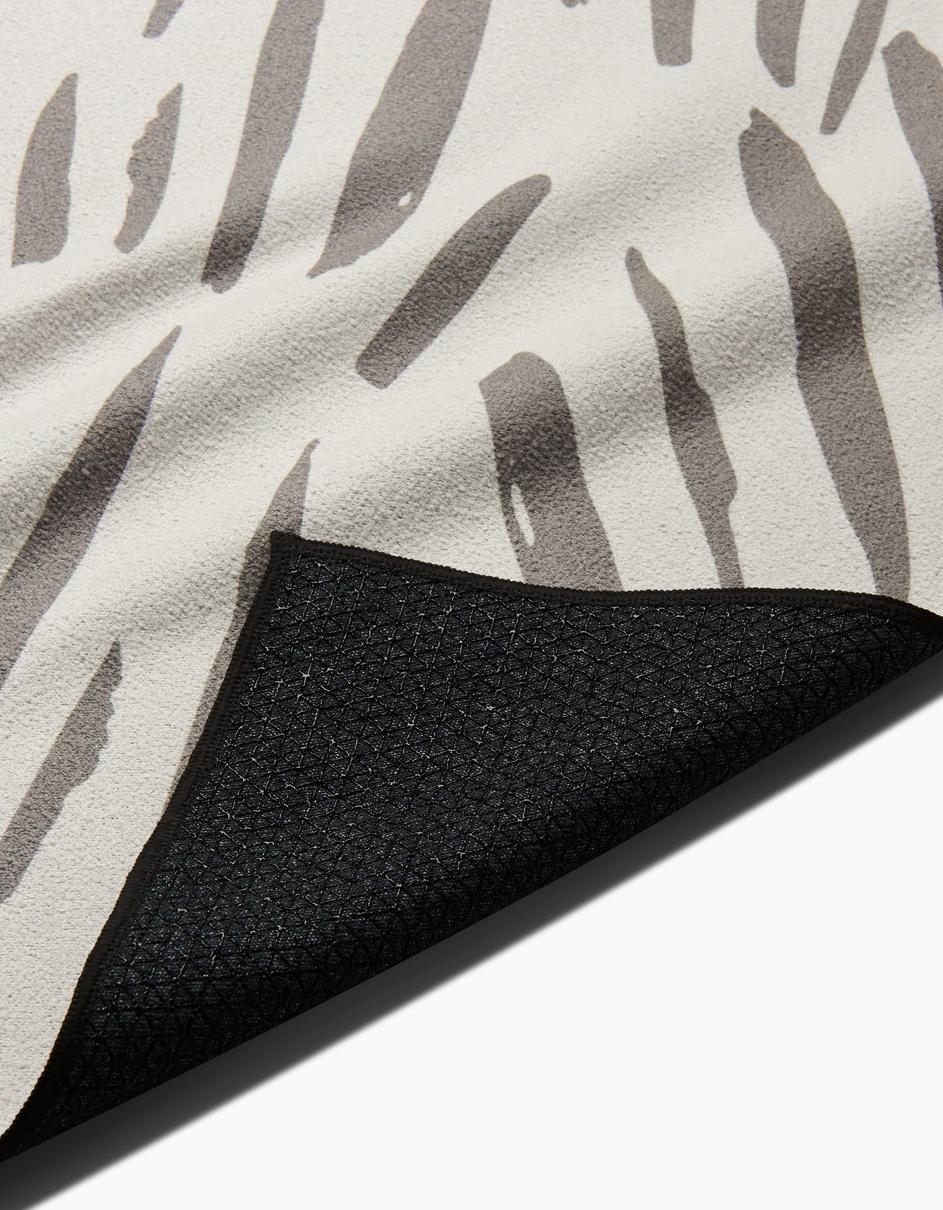 A close-up of the Hufton Studio Country Fields slip-resistant yoga towel featuring a beige and gray abstract pattern, with one corner lifted to reveal its textured black underside.