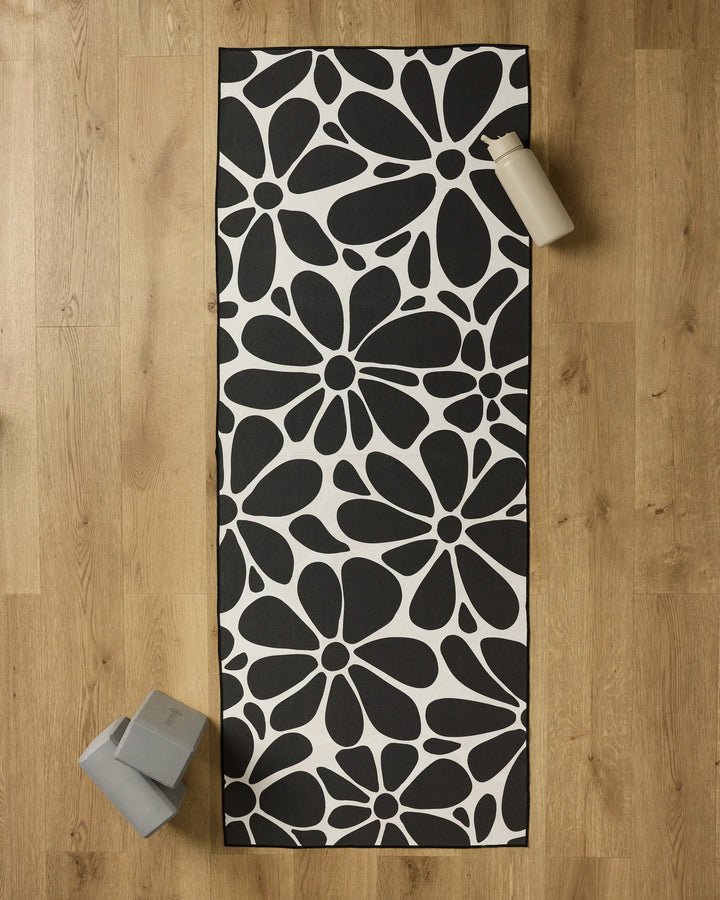 The GEOMETRY Cute Flowers yoga mat with a bold black and white floral pattern is laid on a wooden floor, topped with a non-slip towel. A water bottle and two yoga blocks sit near the mat’s corners.