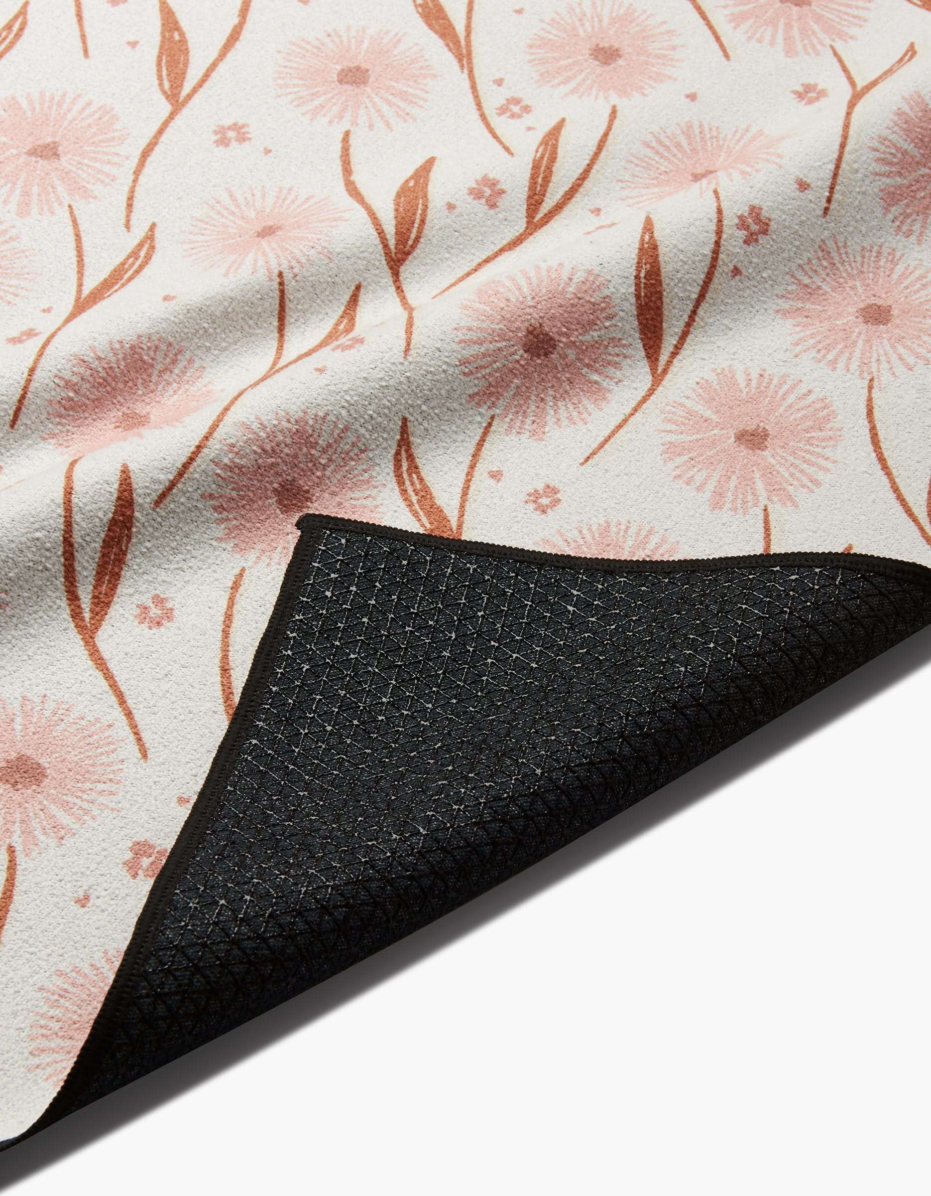 A close-up of the Hufton Studio Daisy Pop yoga towel shows its white surface with a pink and brown floral design and a partially folded black slip-resistant backing revealing the underside.
